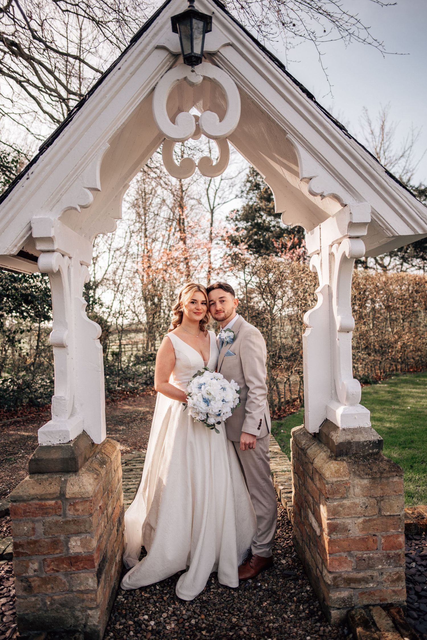 Bride and groom stand under ornate white arch with bouquet at wedding venue.