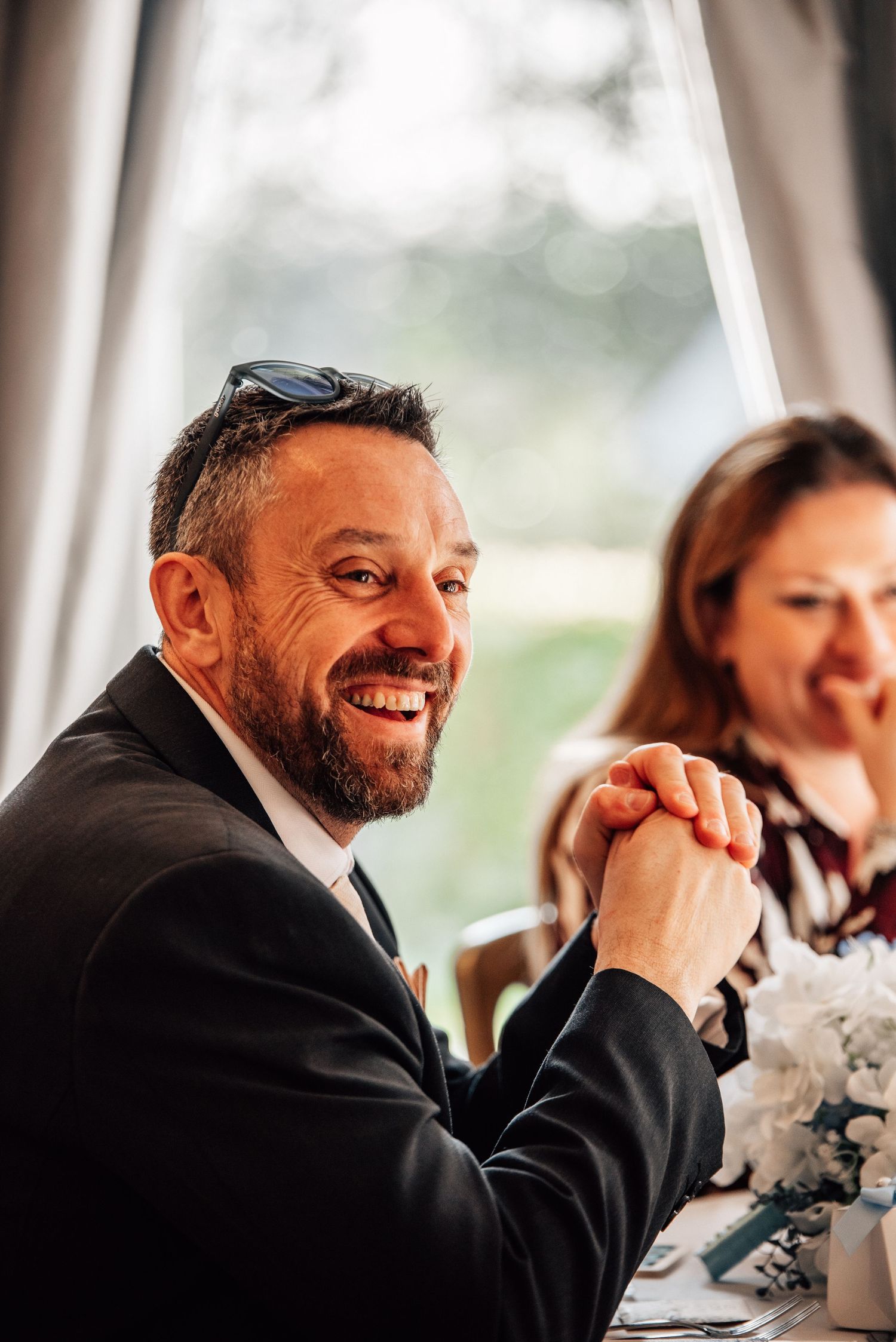 Smiling bearded man in black suit with sunglasses on head, sitting at a wedding reception table.