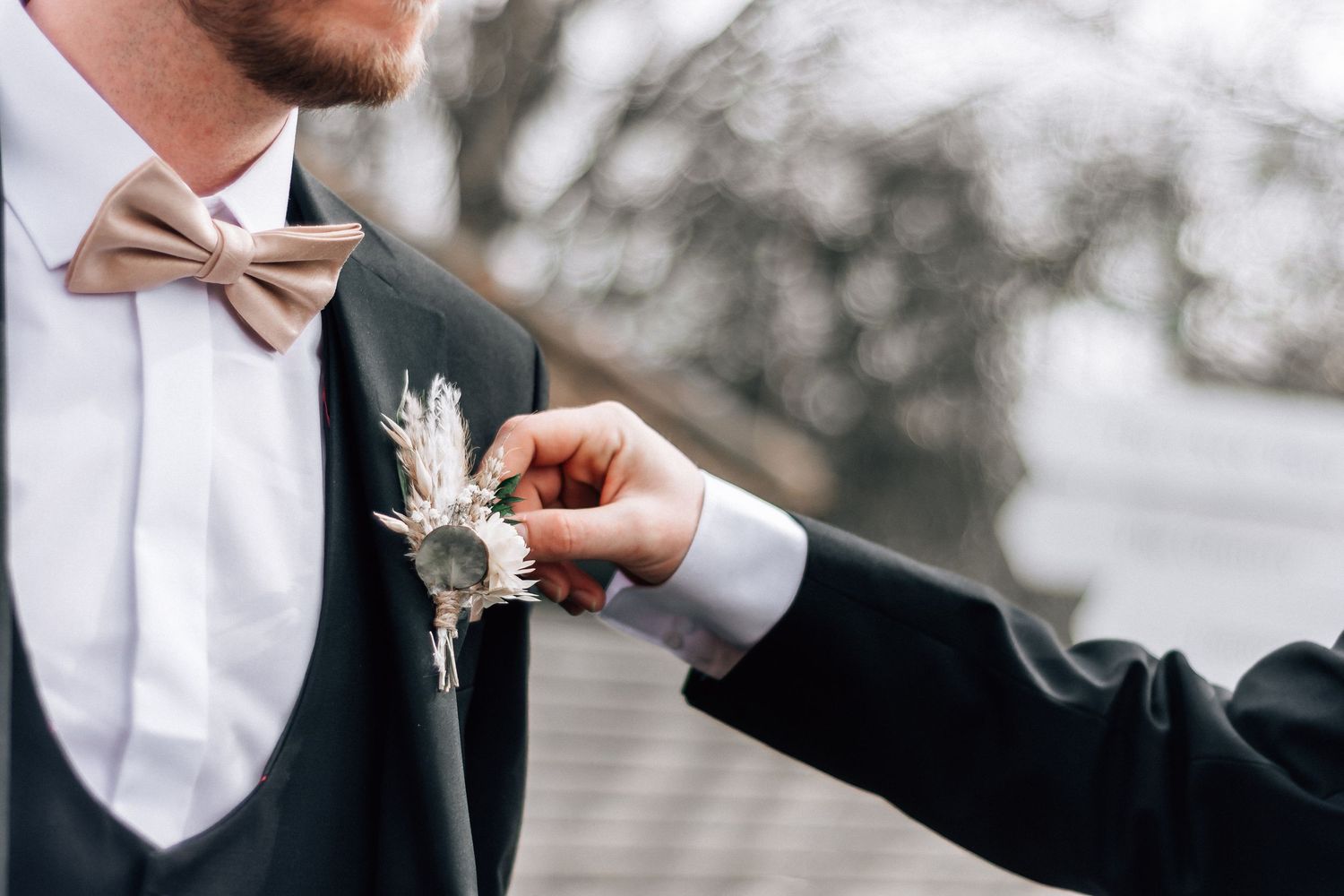 A groom in a black tuxedo with bow tie gets a floral boutonniere pinned to his lapel at a wedding.