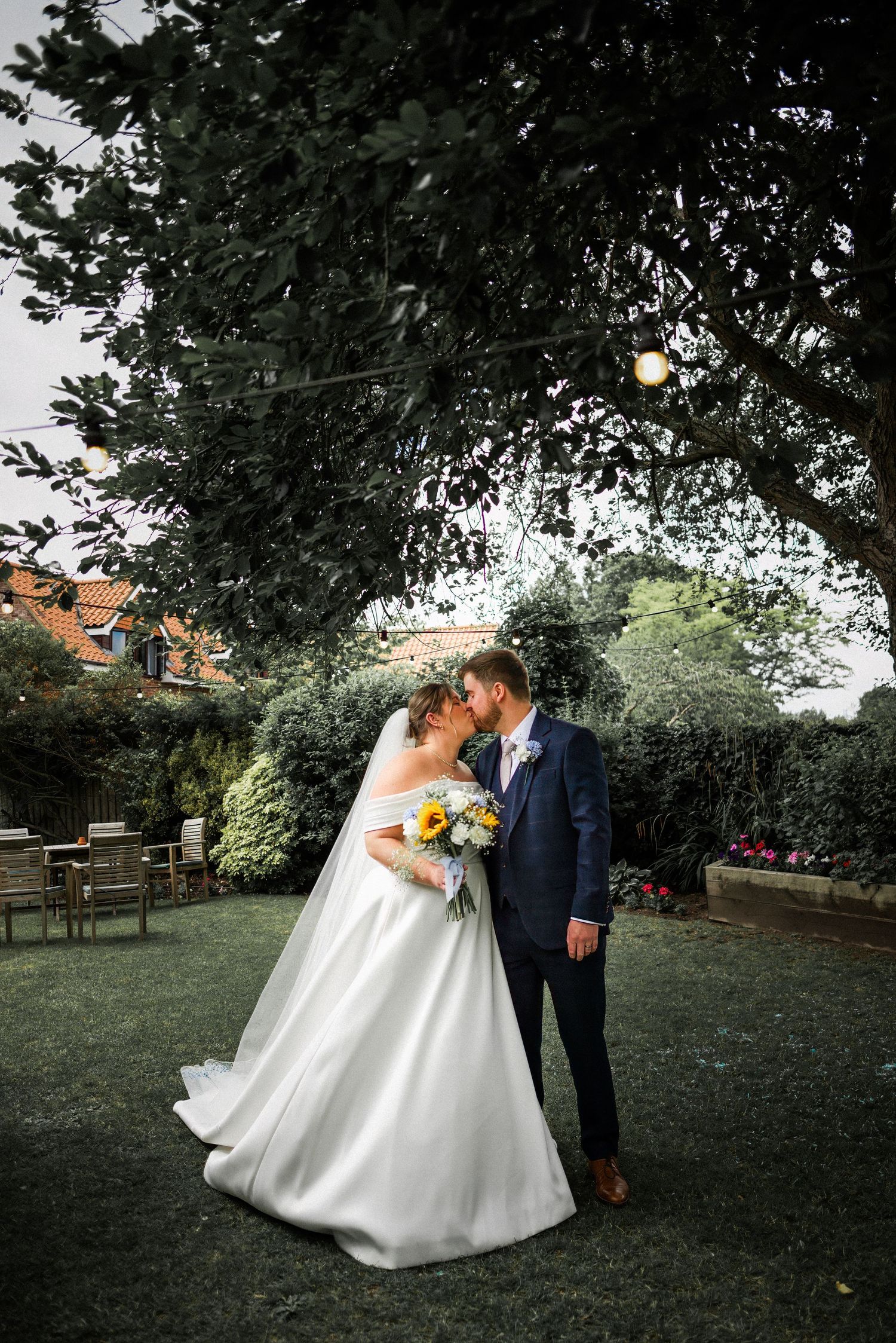 Bride and groom share a romantic kiss in a lush garden, bride in white gown holding sunflower bouquet, groom in navy suit.