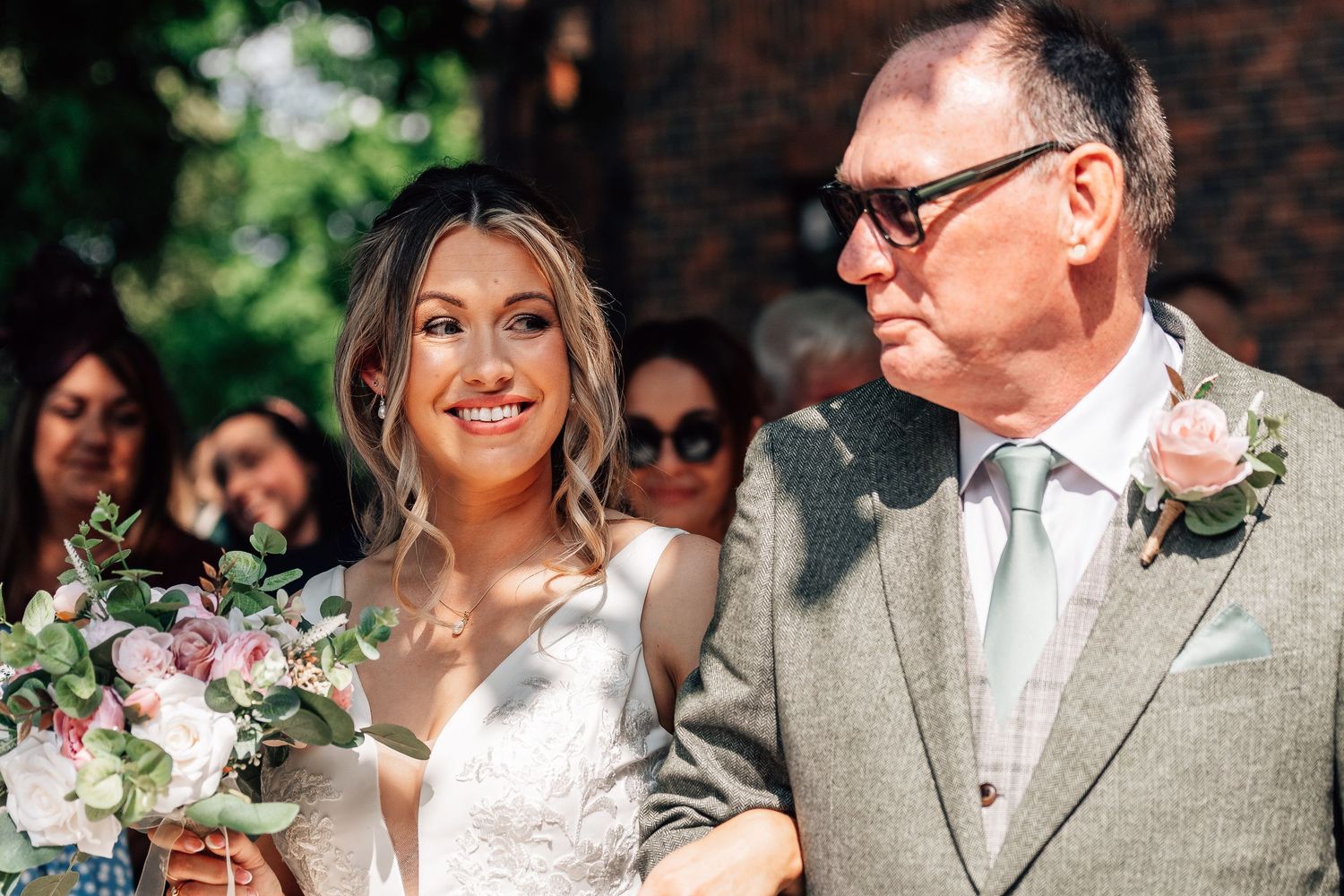 Bride smiling with bouquet beside older man in suit at outdoor wedding ceremony.