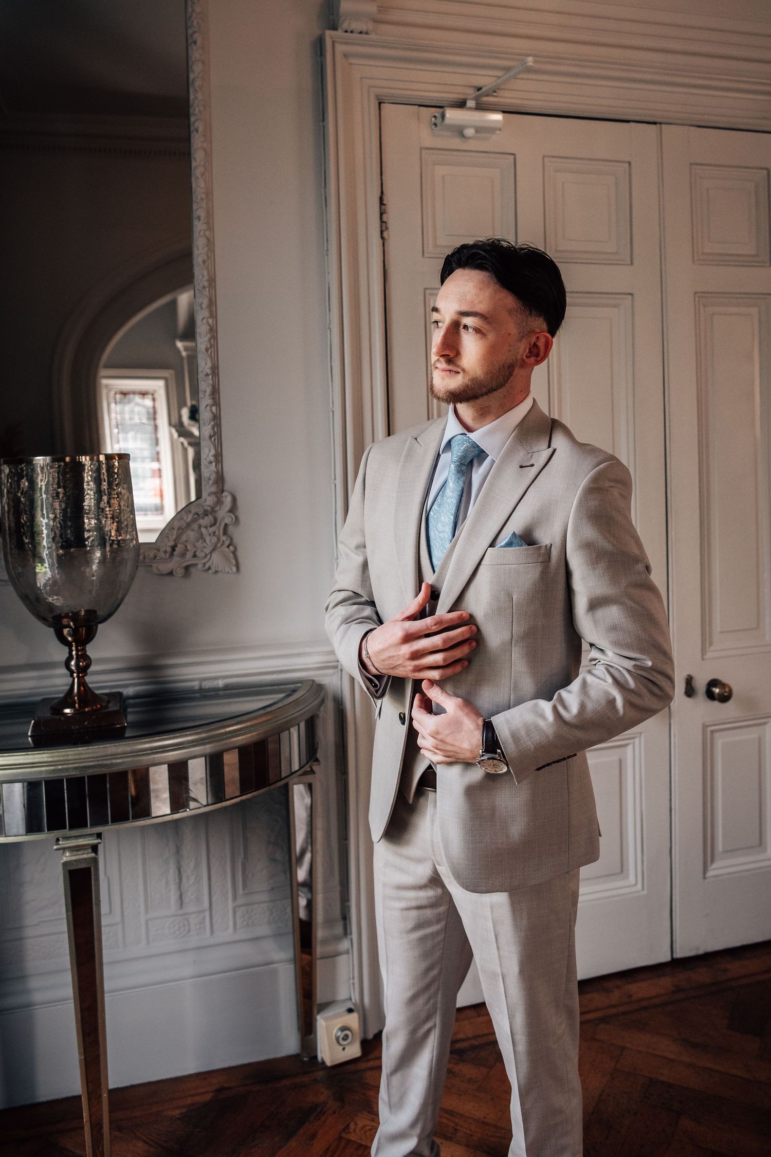 Handsome groom in a beige suit and blue tie standing confidently in a classic interior.