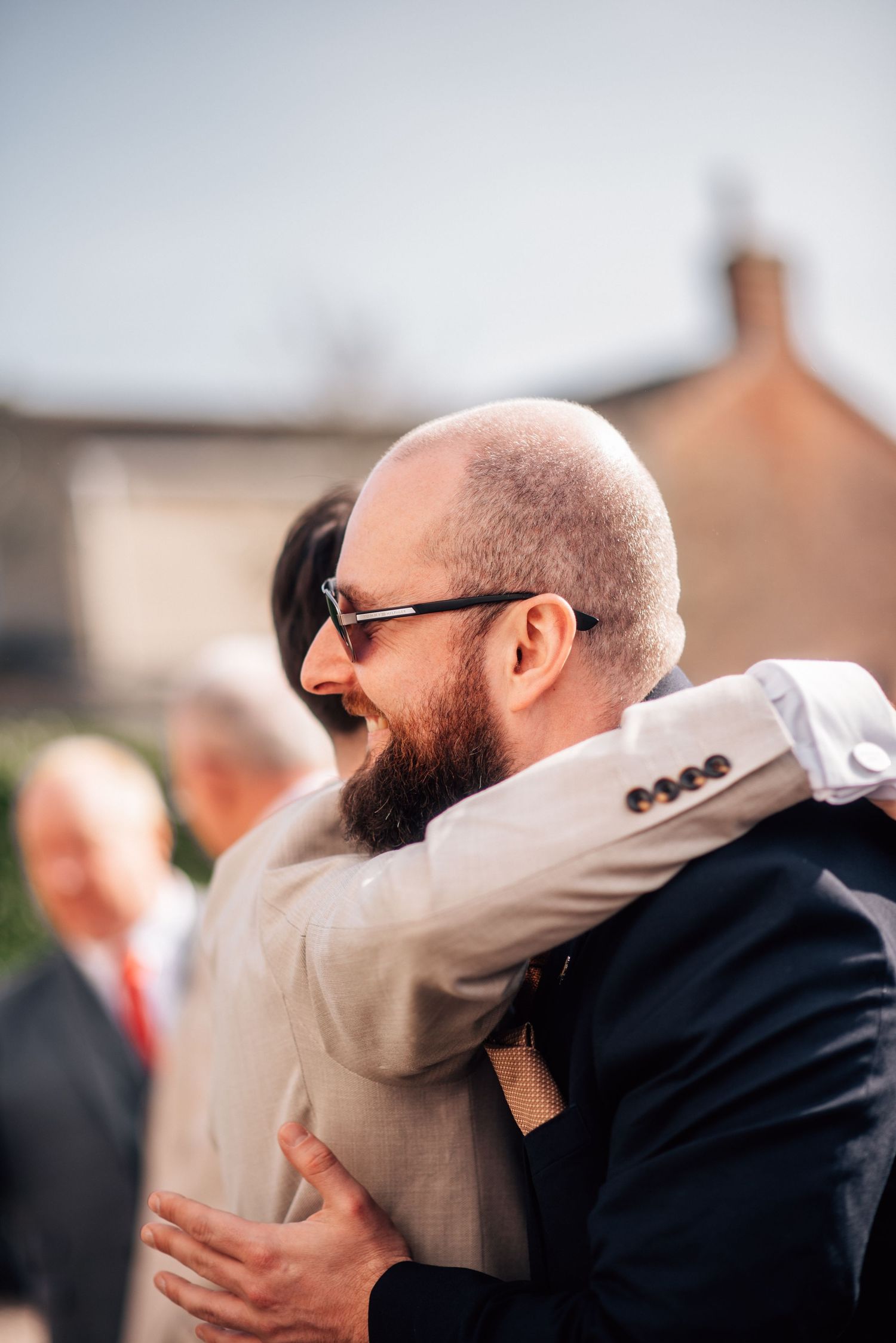 Bald bearded man wearing sunglasses laughing while embracing someone at an outdoor wedding celebration.