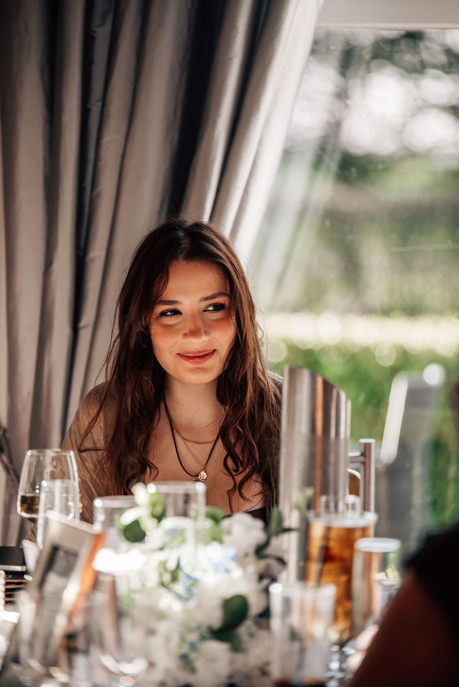 Young woman with long brown hair smiling at a elegantly set dining table with glassware and white floral arrangements.