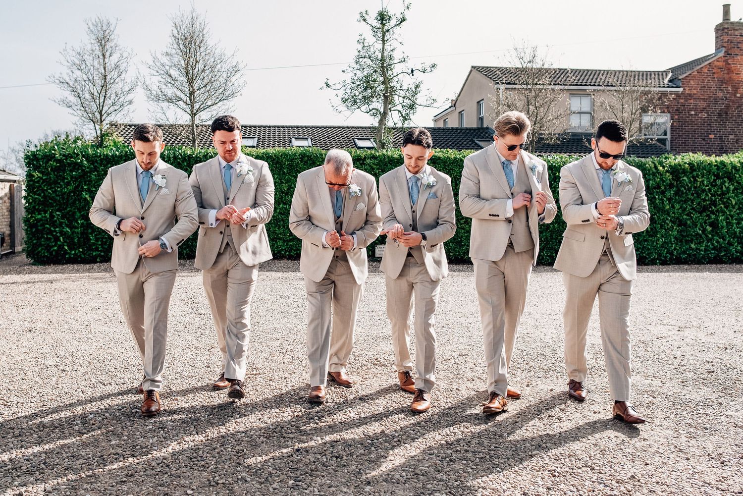 Six groomsmen in matching tan suits walking together outdoors at a wedding venue on a sunny day.