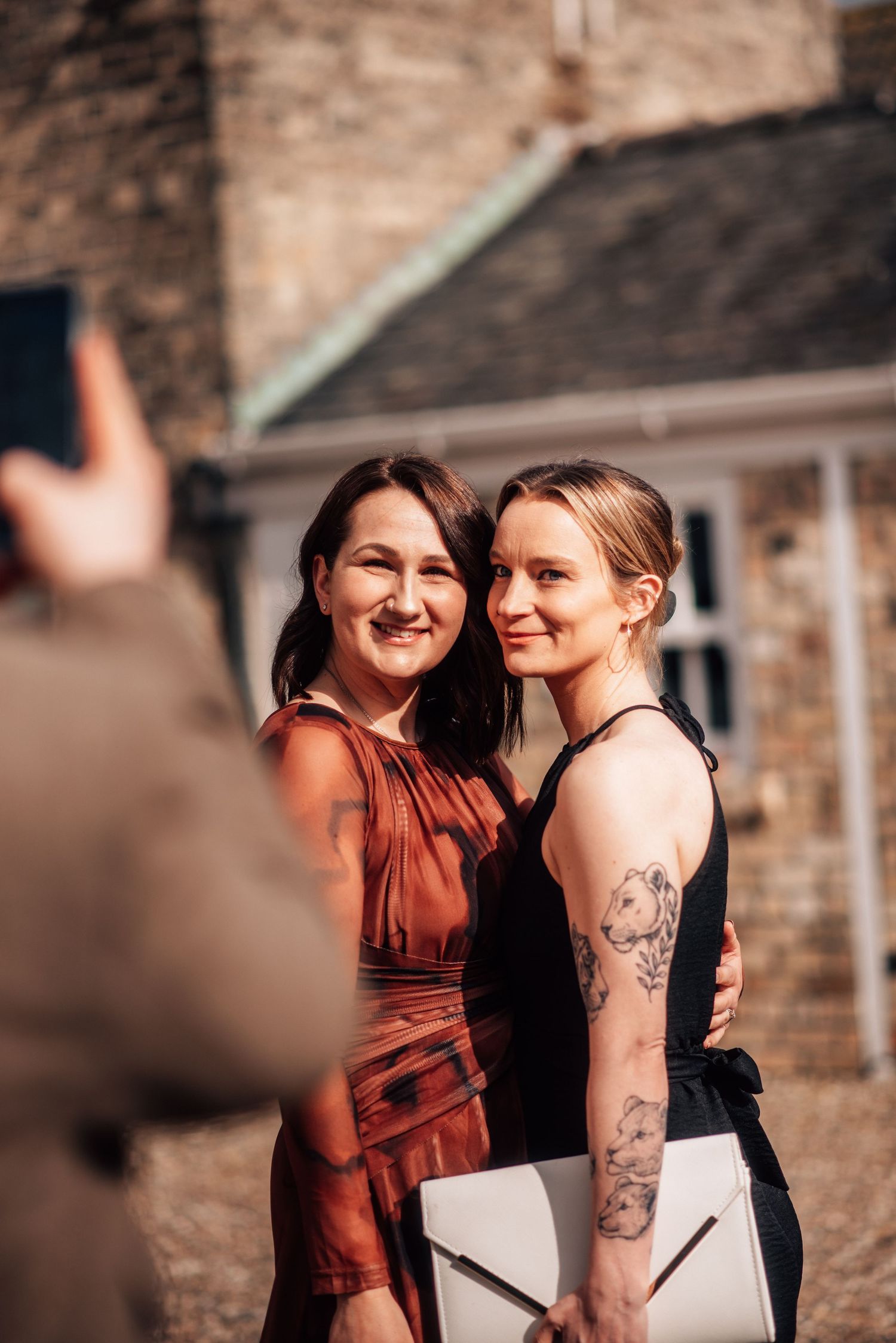 Two women in elegant dresses pose together smiling outdoors at a wedding event.