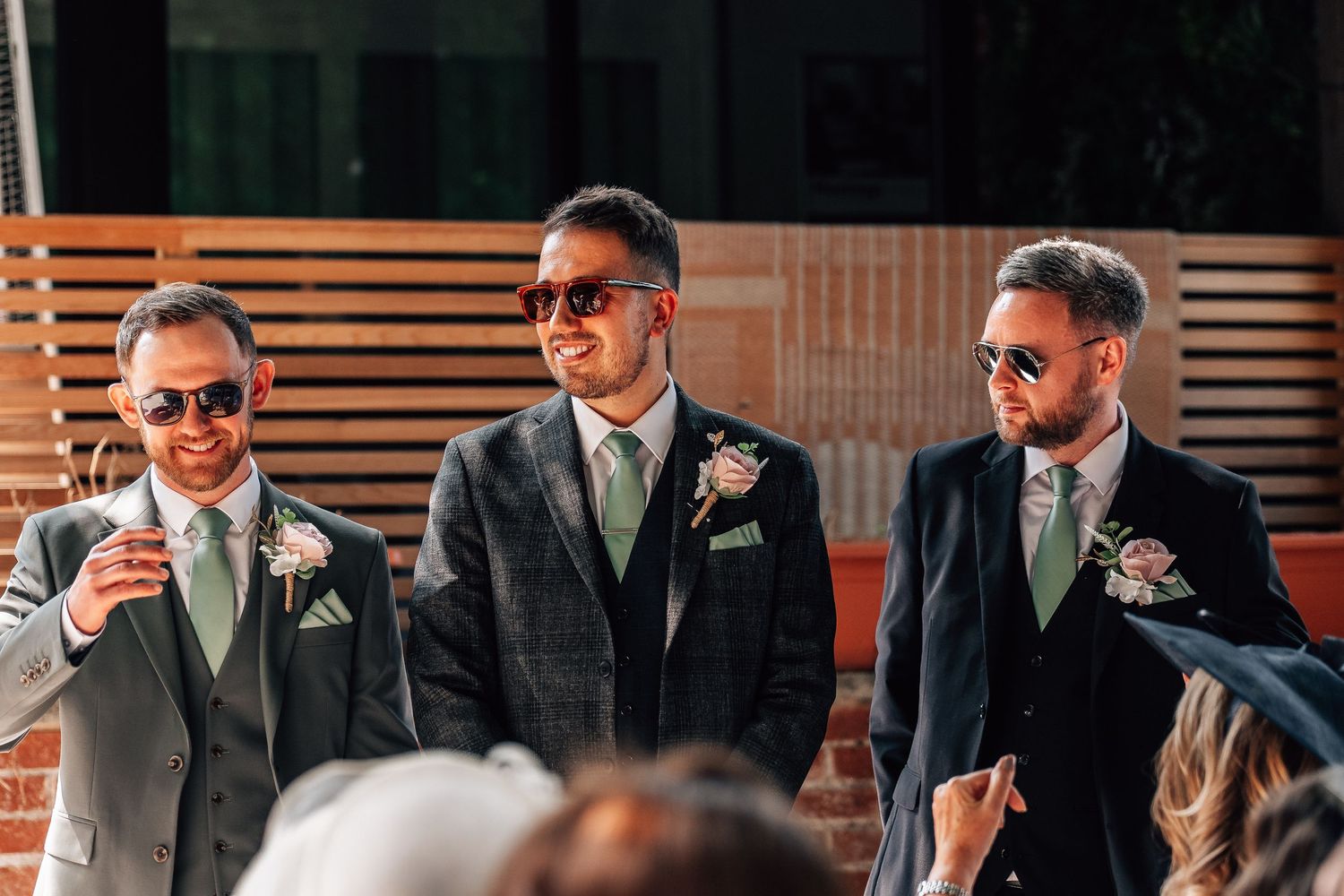 Three groomsmen in dark suits with sage green ties and floral boutonnieres smiling at an outdoor wedding ceremony.