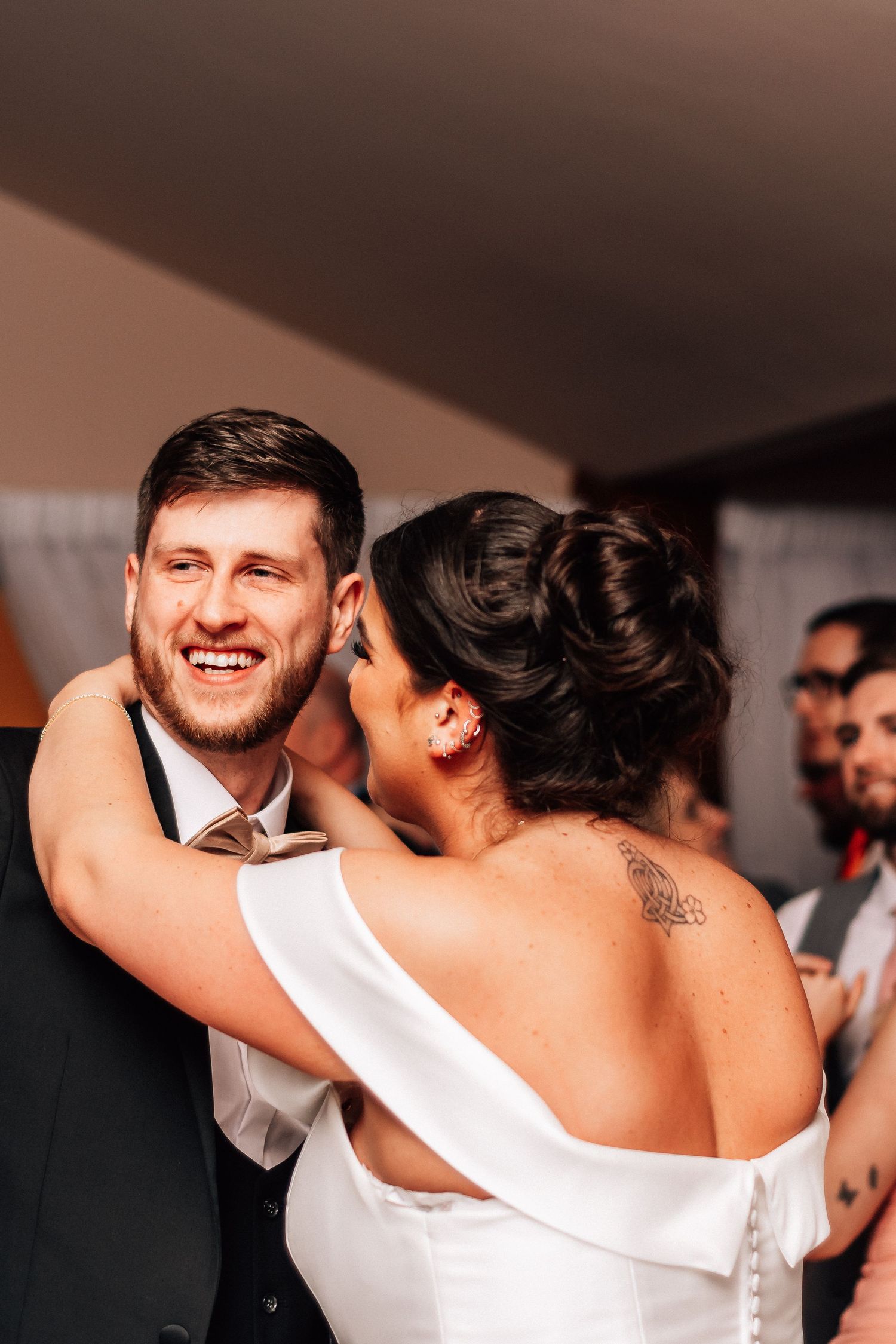 Smiling groom in black suit shares first dance with bride in white off-shoulder gown at wedding reception.