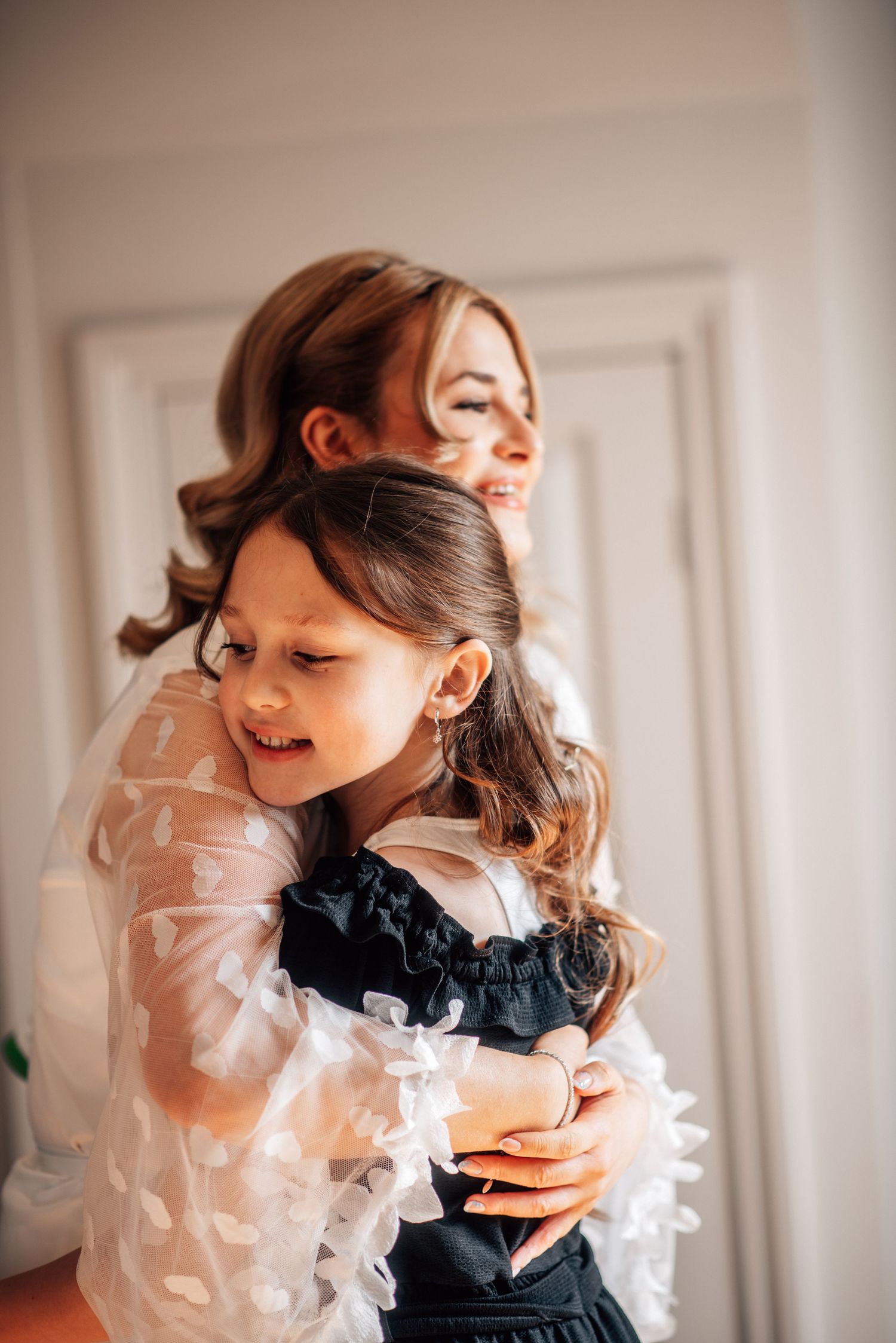 A bride in a white tulle dress lovingly embraces a young girl in a black dress, both smiling joyfully.