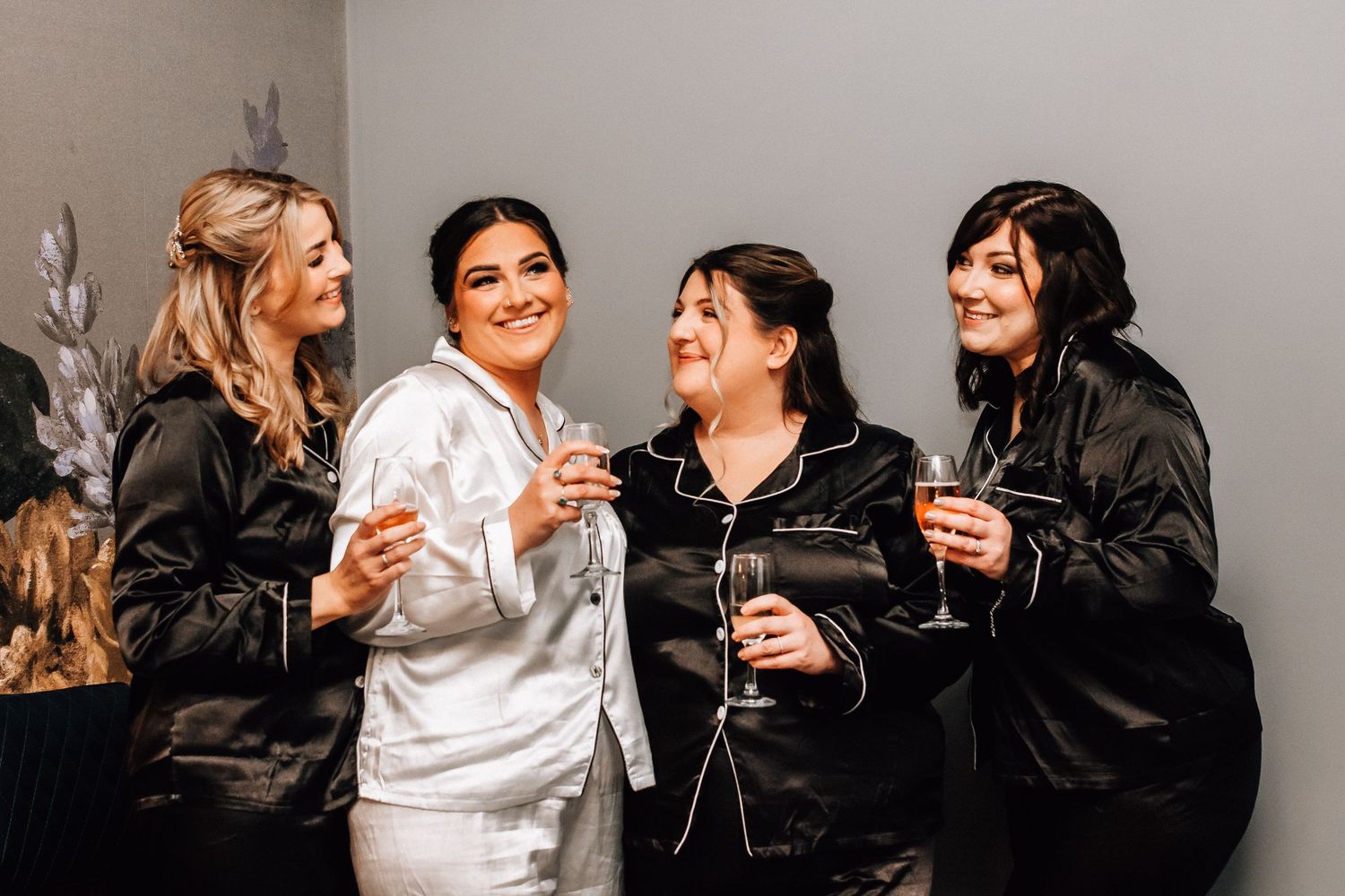 Bride in white satin robe celebrating with bridesmaids in black robes, toasting champagne glasses before the wedding.