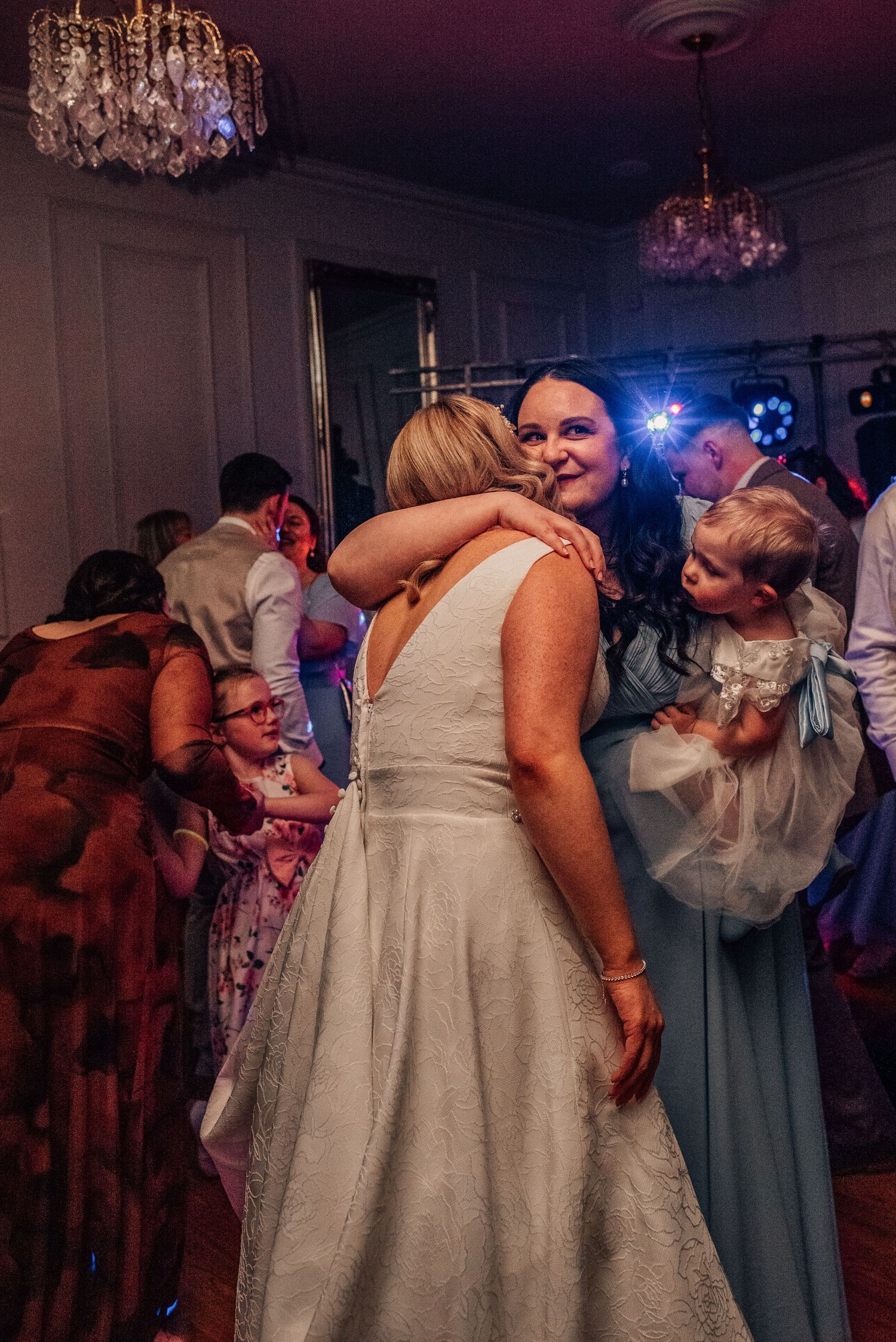 Bride in white lace gown hugging guest in blue dress at wedding reception with chandeliers and dance floor.