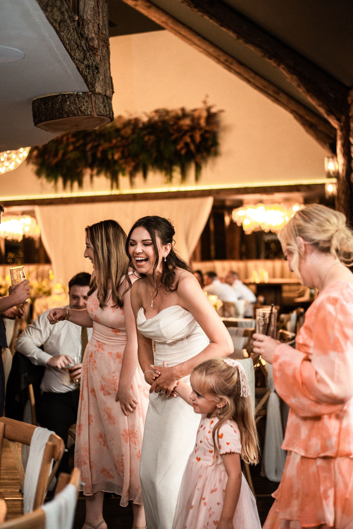A laughing bride in a white strapless gown dances joyfully with guests at a rustic wedding reception.