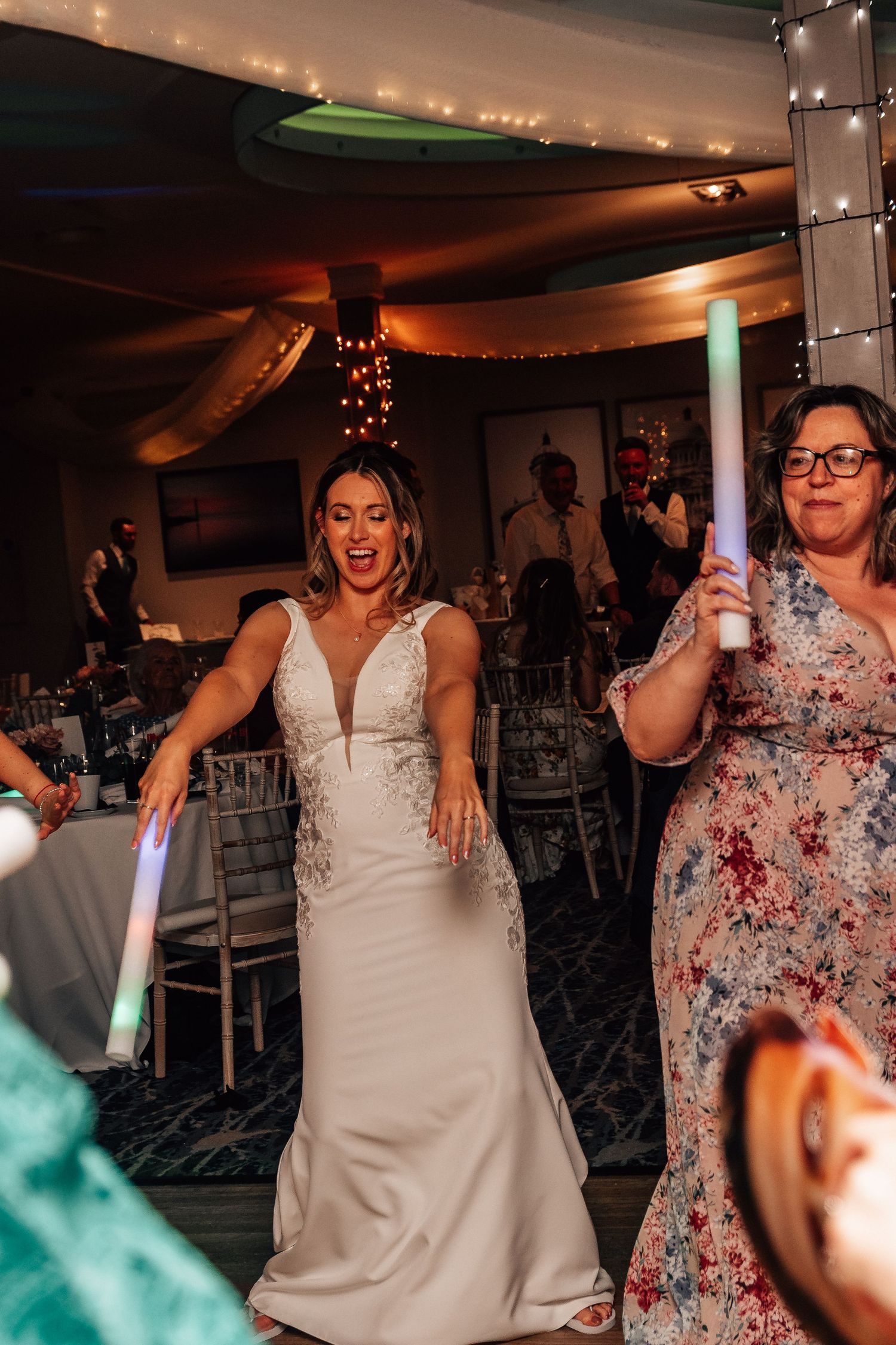 Excited bride in a white gown dancing energetically with guests holding glow sticks at her wedding.