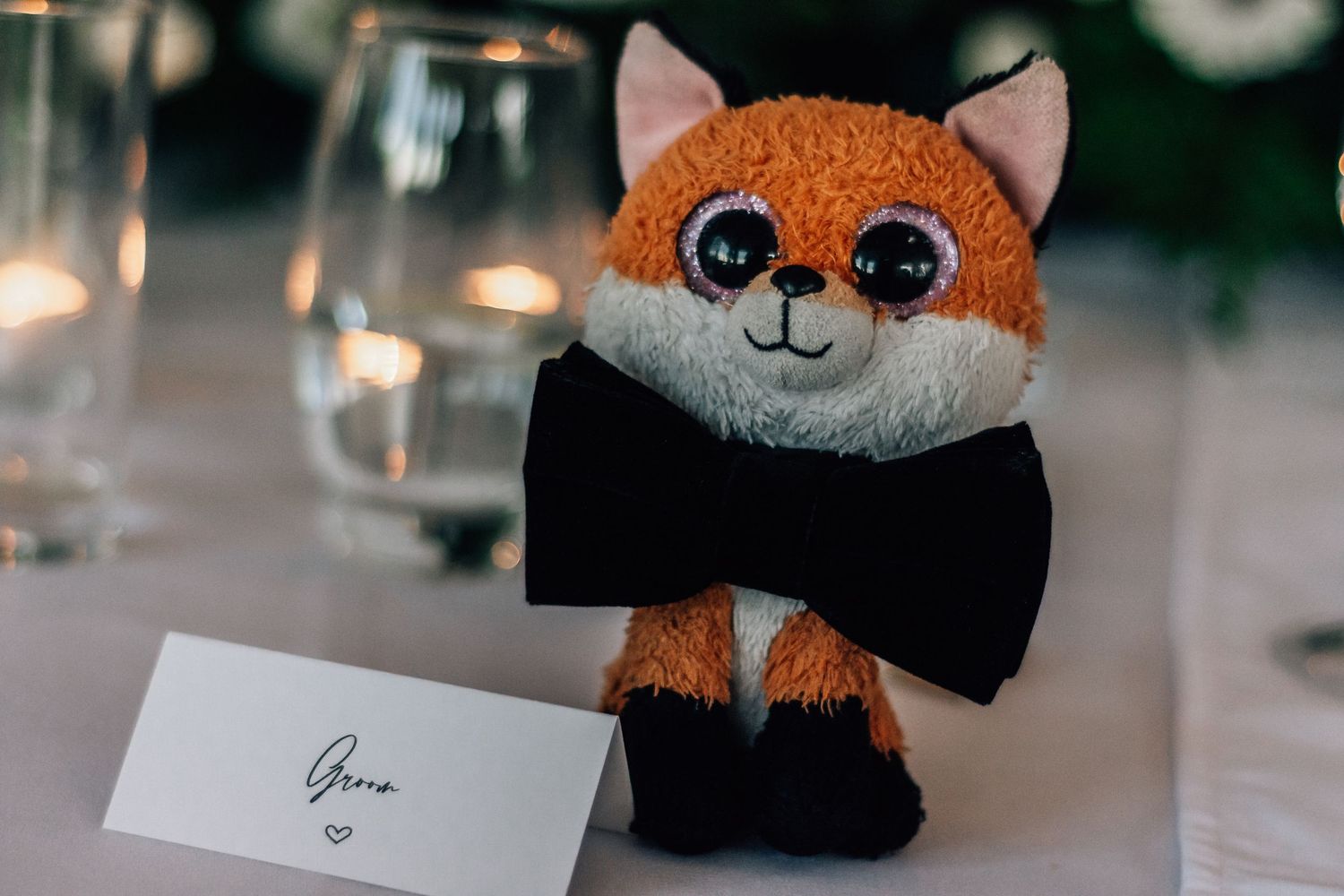 A cute orange fox stuffed animal wearing a black bow tie sits next to a wedding place card on a table.