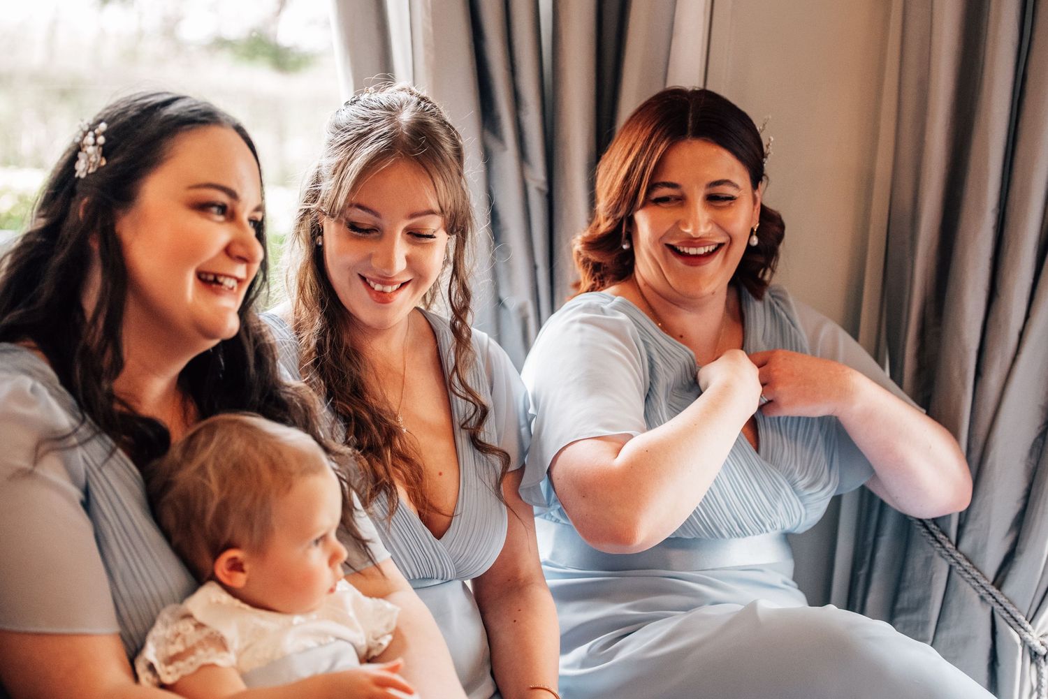 Three bridesmaids in light blue dresses laugh together, one holding a baby, in a bright wedding preparation setting.