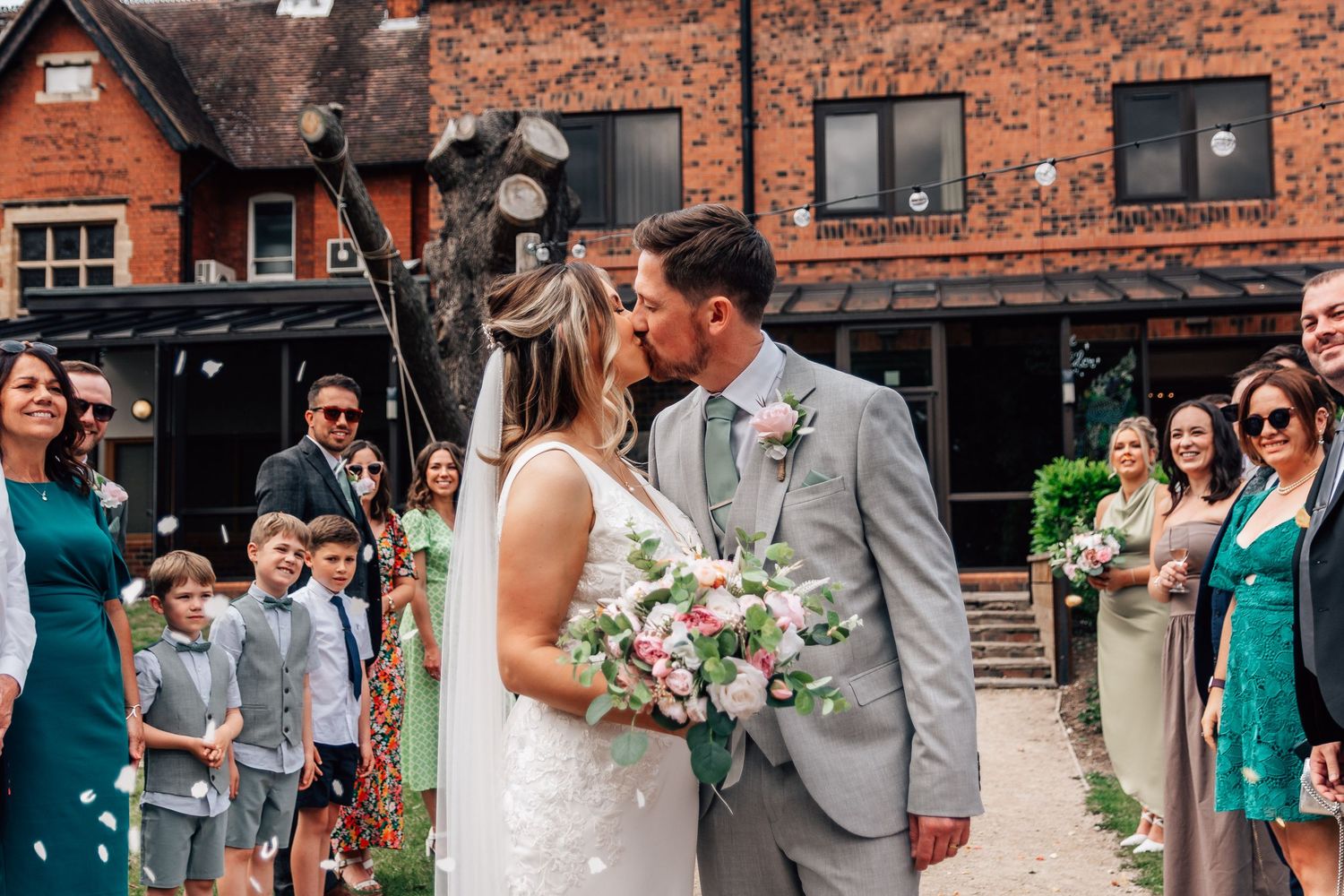 Bride and groom share a kiss surrounded by guests throwing confetti outside a rustic brick wedding venue.