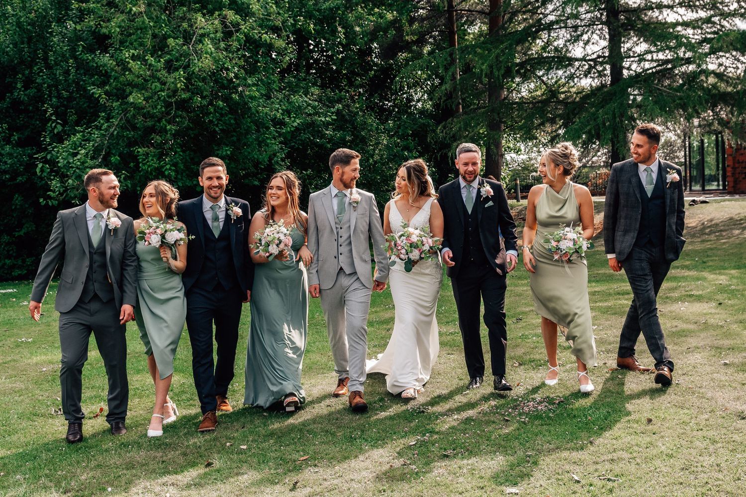 Wedding party walking outdoors on grass, bride in white gown, bridesmaids in sage green dresses, groomsmen in suits.