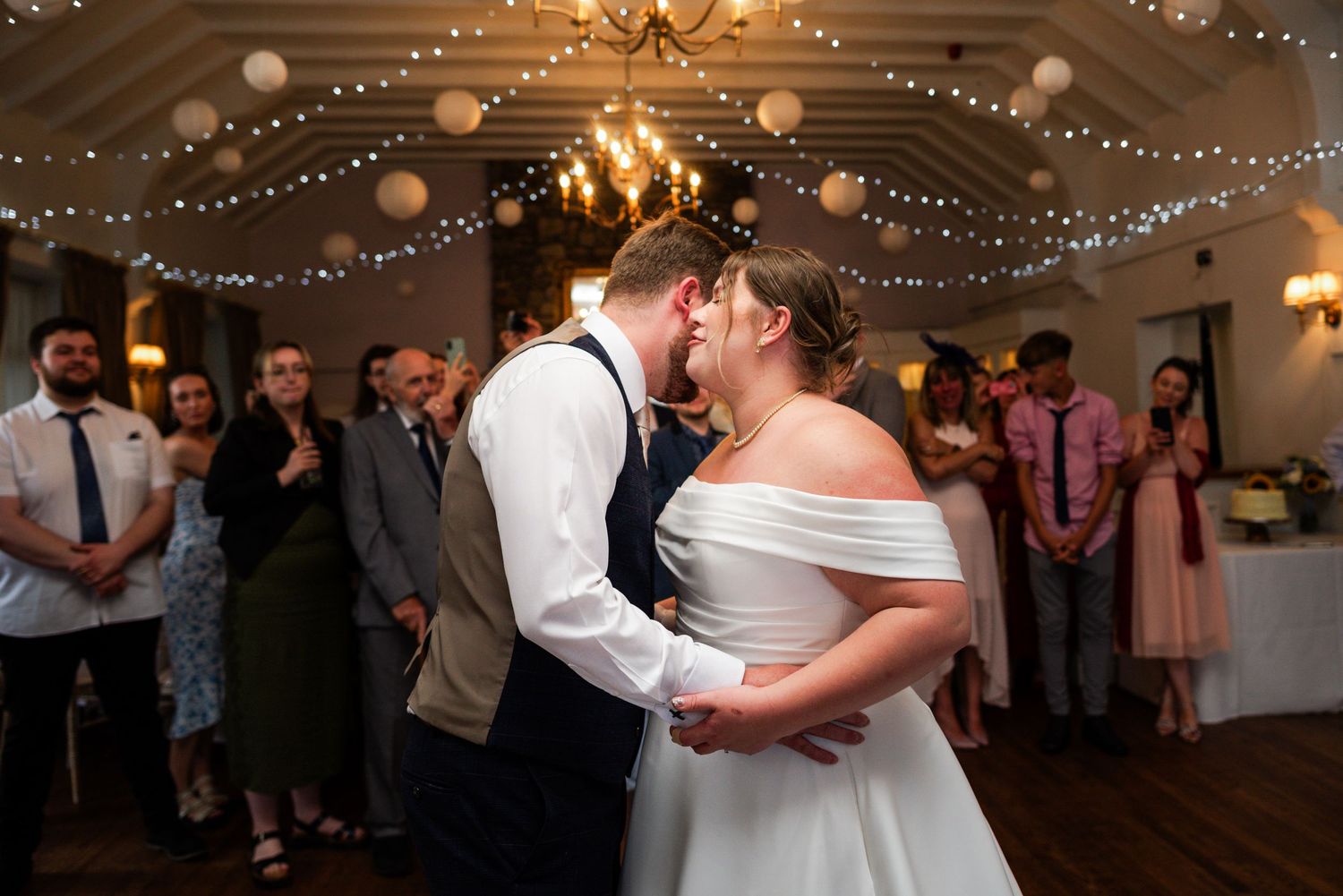 Bride and groom share first dance surrounded by guests in a warmly lit wedding reception venue with fairy lights.