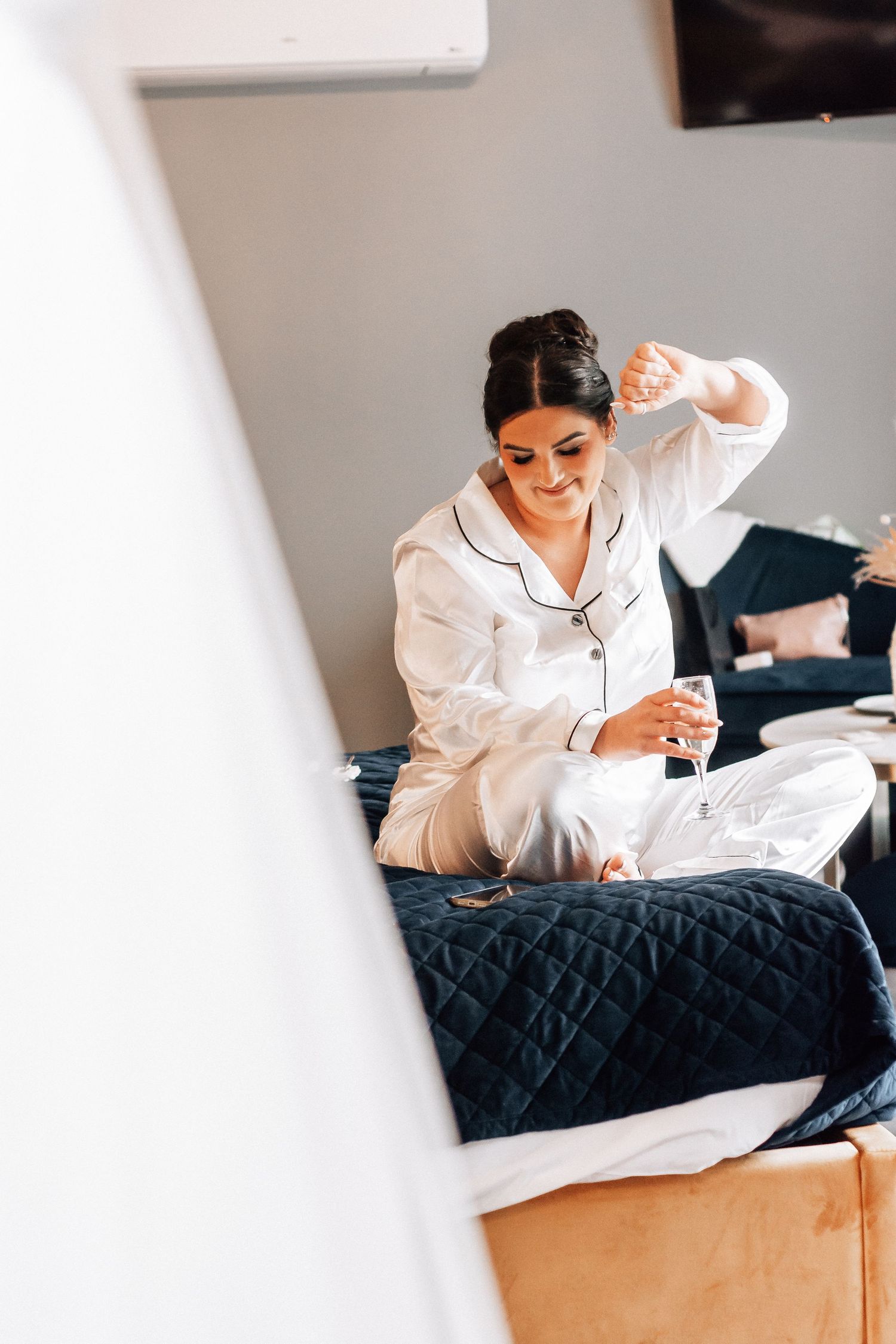 Woman in white pajamas sitting on a navy blue bed, holding a champagne glass in a bright hotel room.