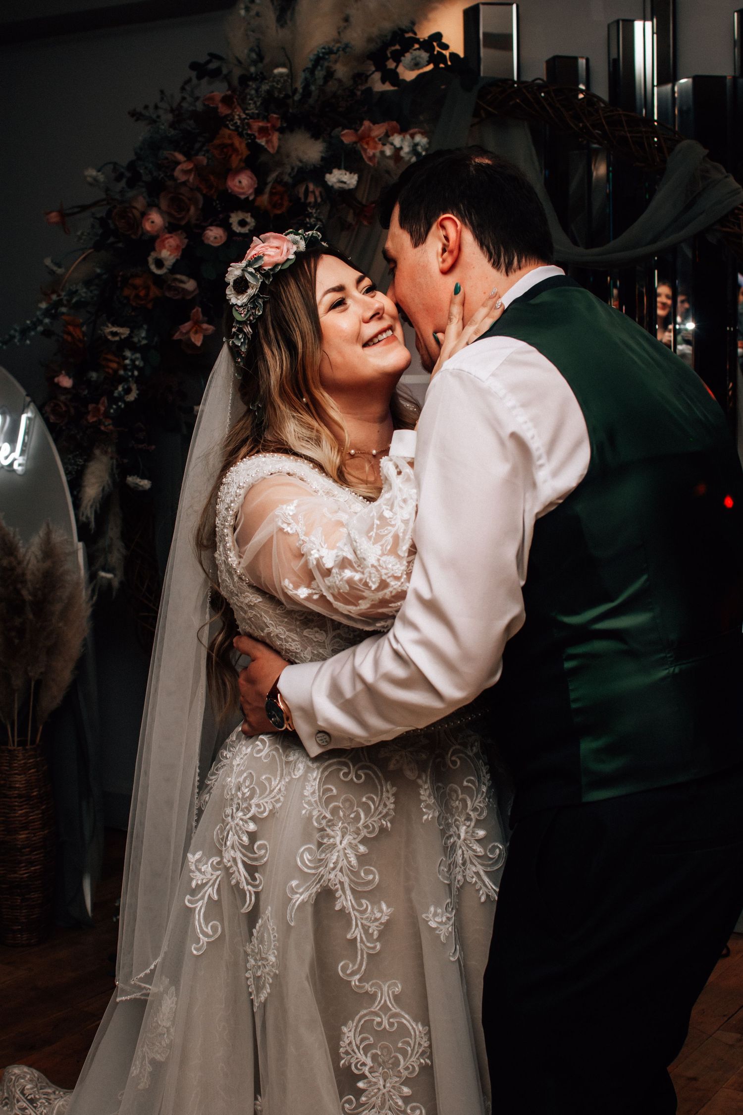 Bride in lace gown and floral crown shares a tender embrace with groom in green vest at romantic wedding reception.