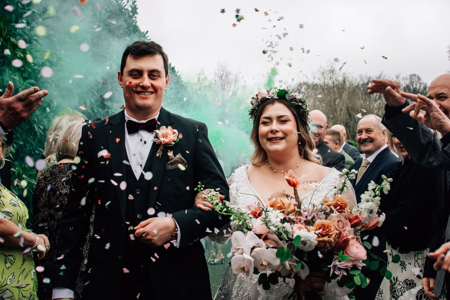Joyful newlyweds walk through confetti and green smoke at their outdoor wedding ceremony.
