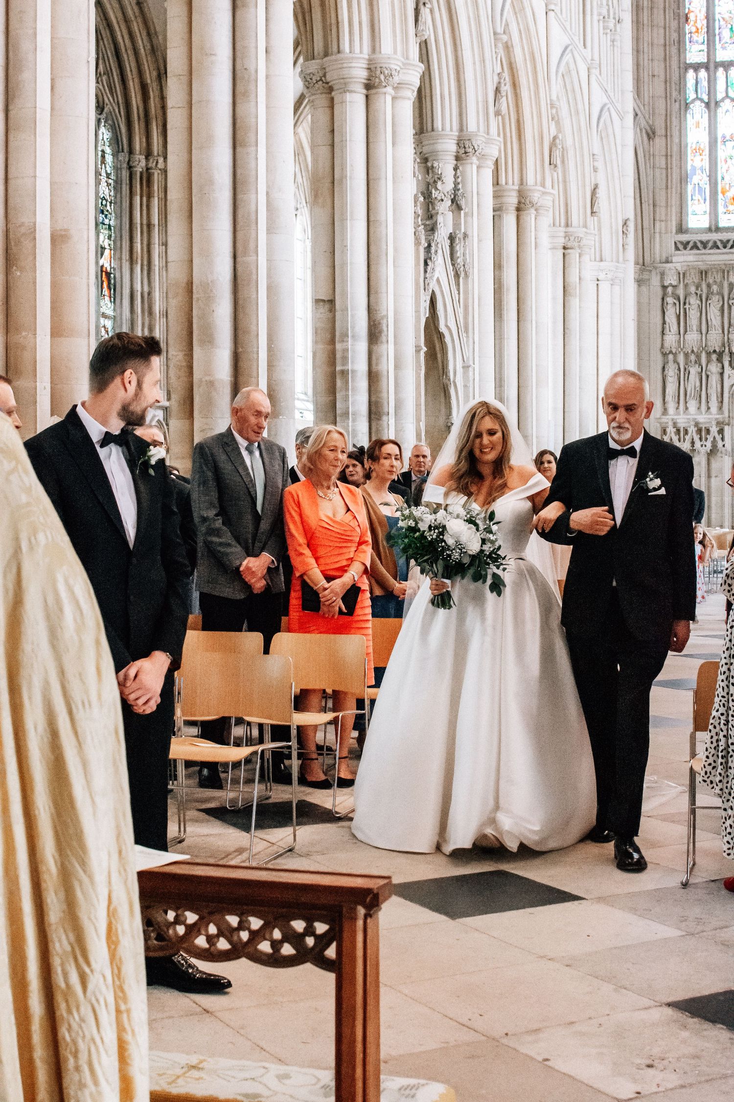 Bride walks down cathedral aisle with father, wearing white ballgown and holding bouquet.