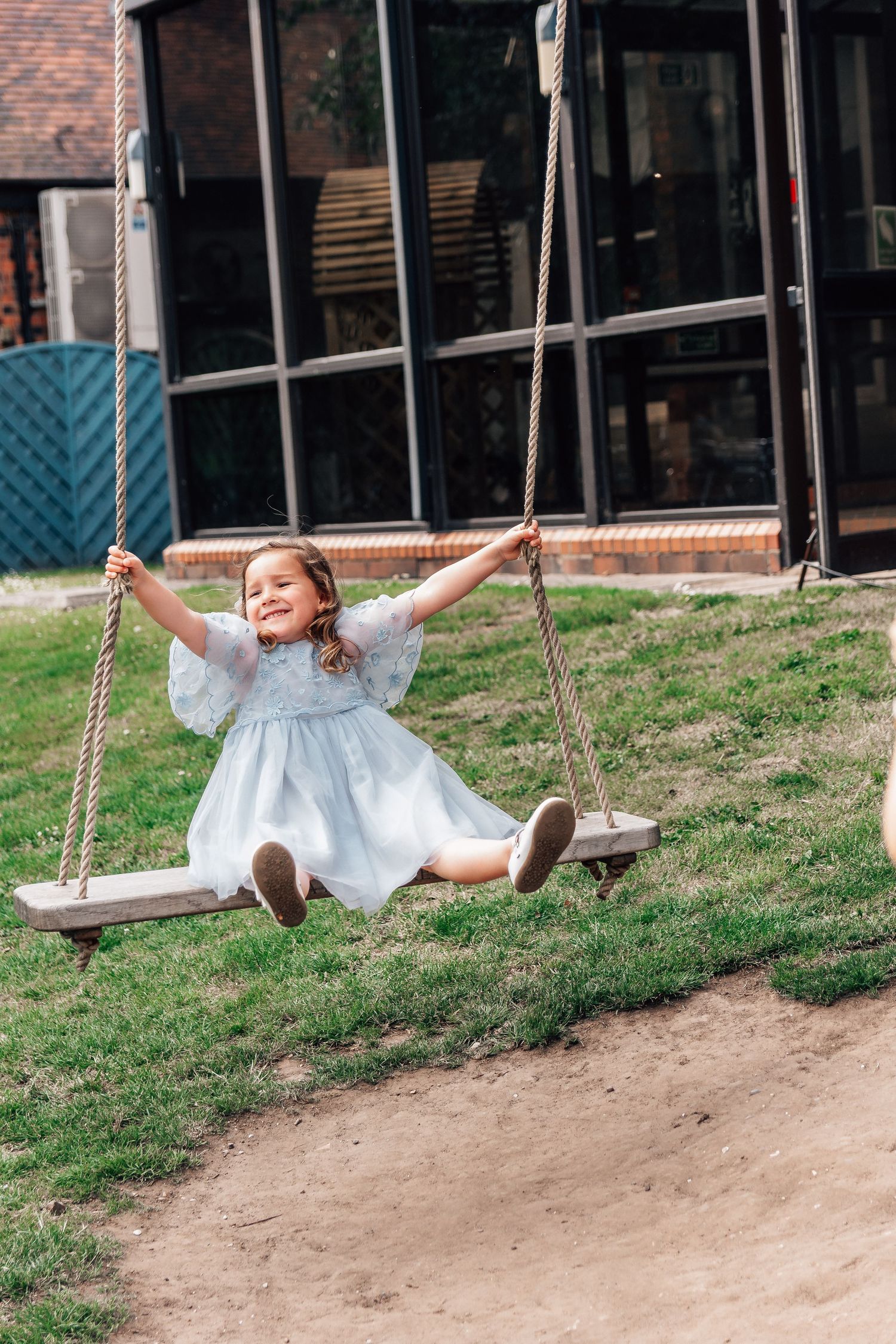 Happy young girl in a light blue dress joyfully swinging on wooden rope swings outdoors.