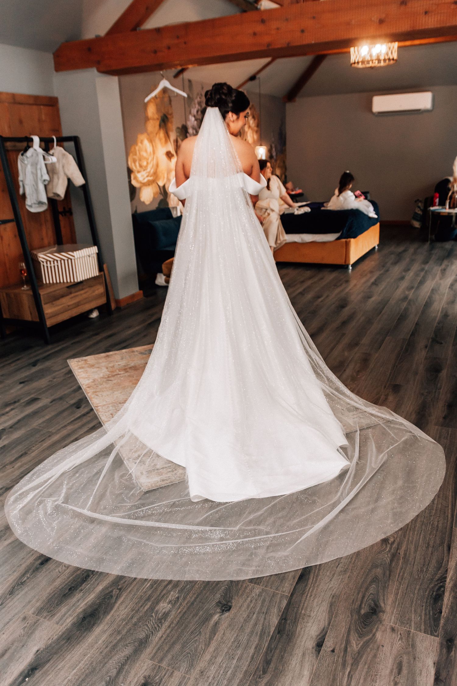Bride in a white wedding gown with a long cathedral veil spread on hardwood floor, viewed from behind in a bridal suite.