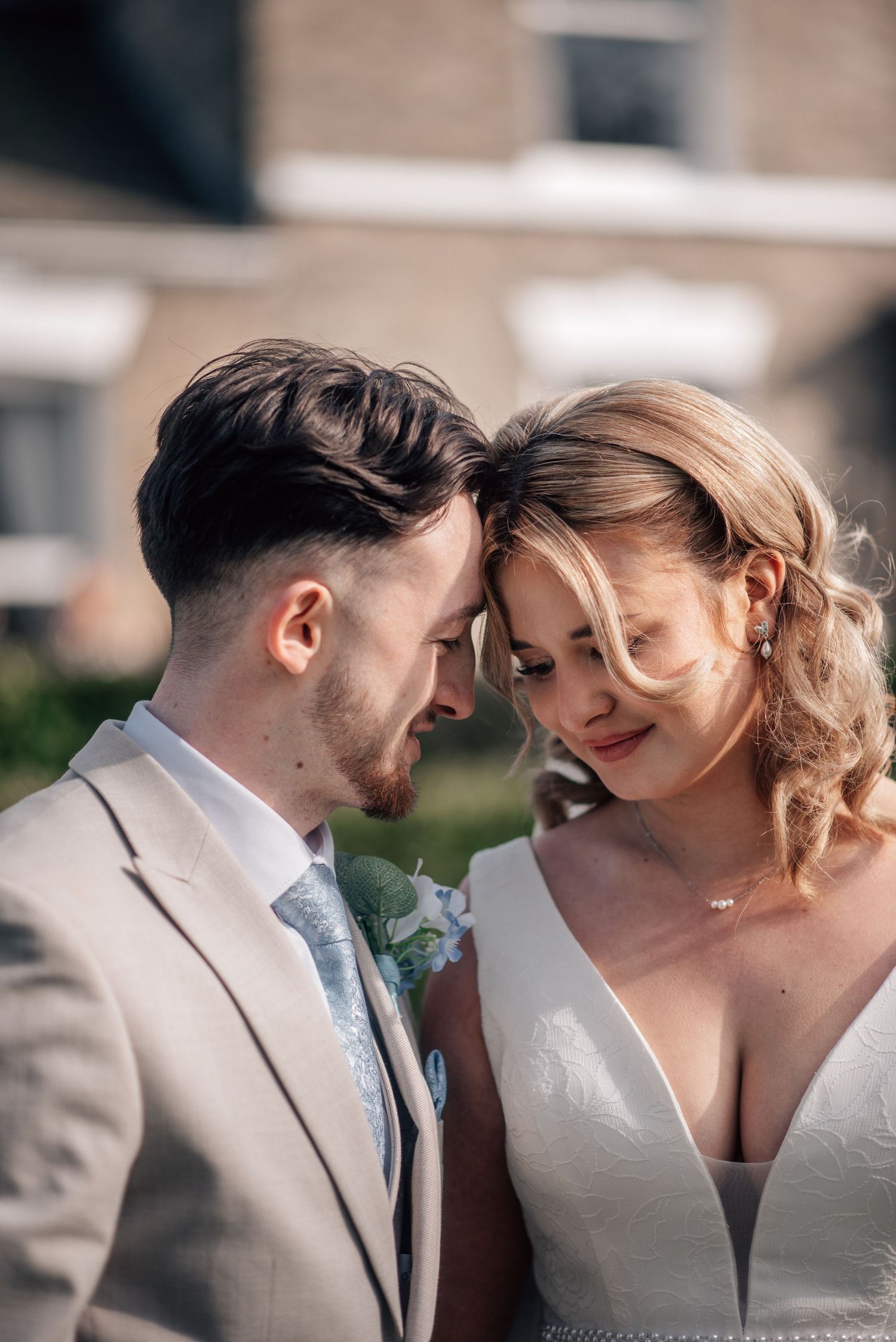 A bride and groom share an intimate forehead touch moment outdoors, wearing wedding attire in warm sunlight.