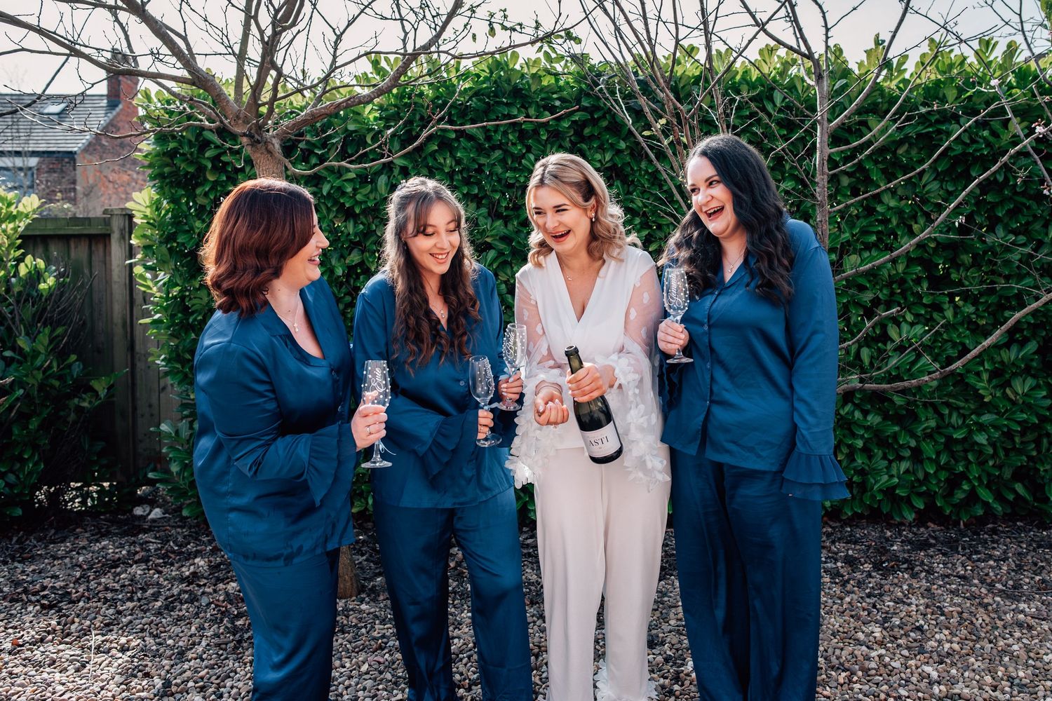 Bride in white pantsuit pops champagne with three bridesmaids in navy suits outdoors.