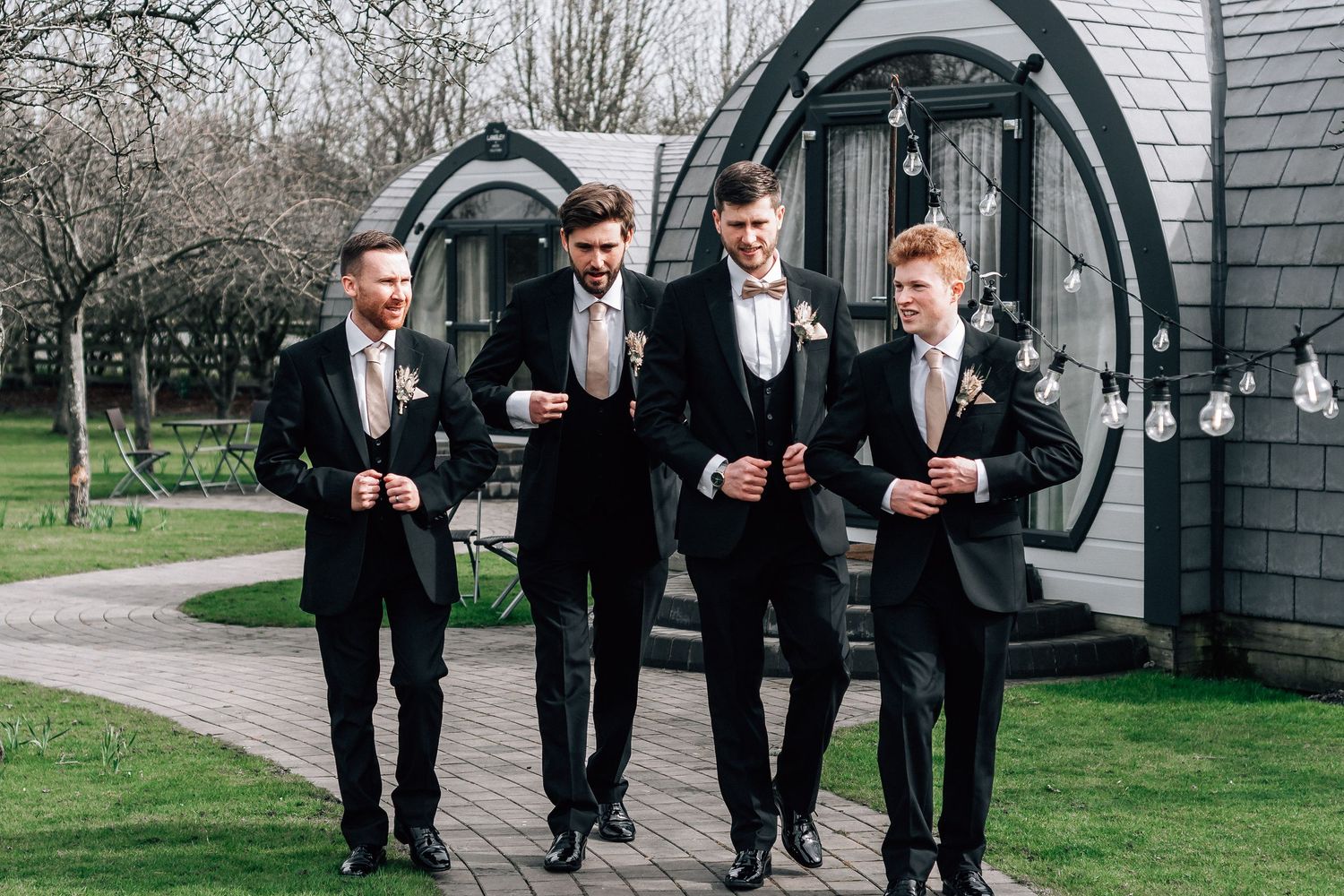 Four groomsmen in black suits with bow ties walking outdoors at a wedding venue.