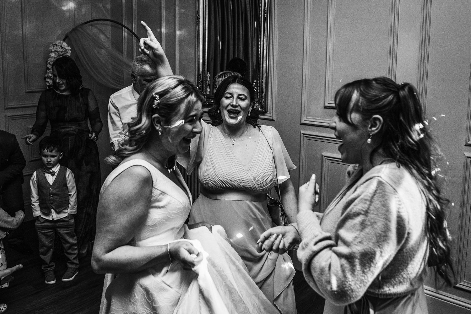 Black and white photo of three women laughing together at a wedding reception, with a young boy visible in the background.