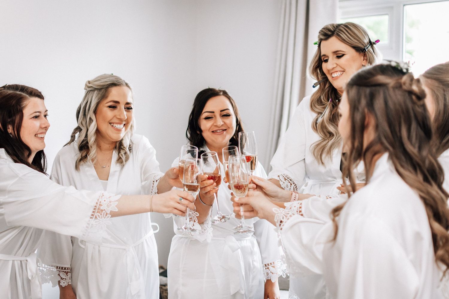 Bridesmaids in white robes toasting with champagne glasses during wedding preparations.