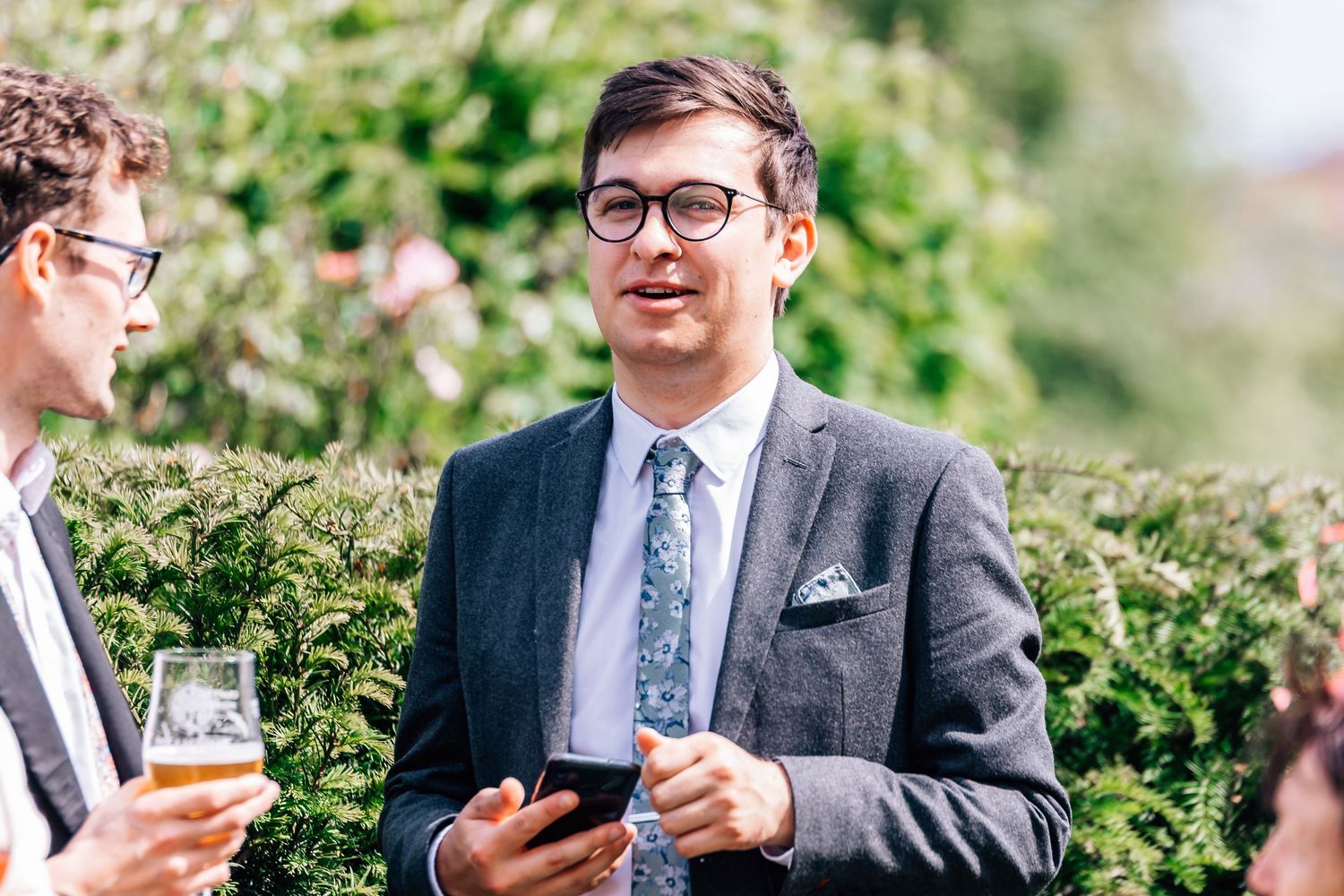 Man in grey suit with floral tie holding smartphone at outdoor wedding reception.