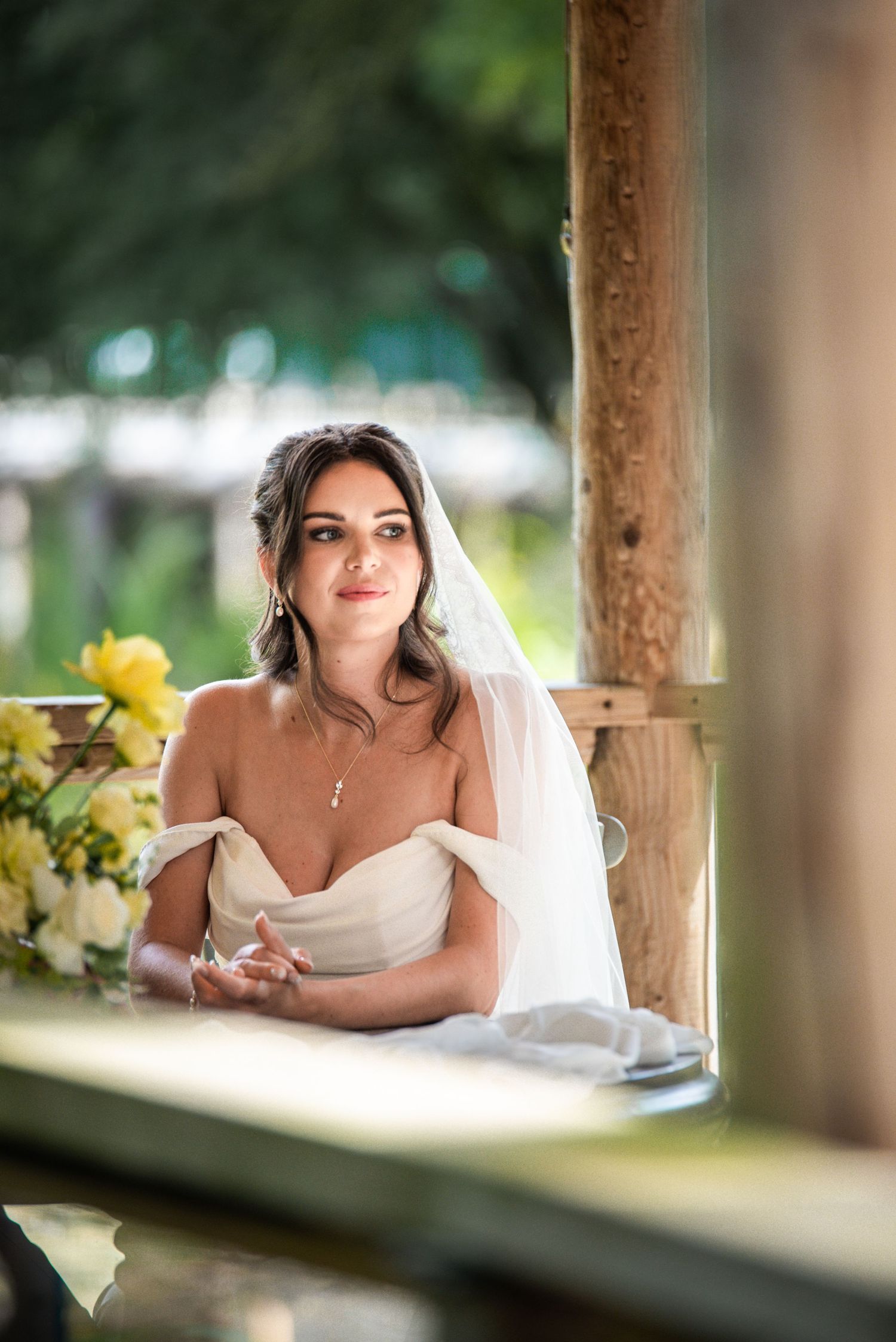 Beautiful bride in white off-shoulder dress and veil seated by rustic wooden post with yellow flowers.