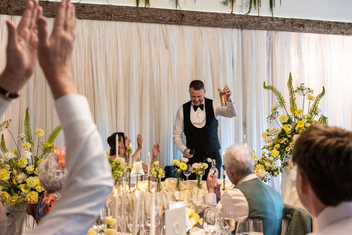 A groom gives a speech at a wedding reception, raising his hand as guests applaud at a floral-decorated table.