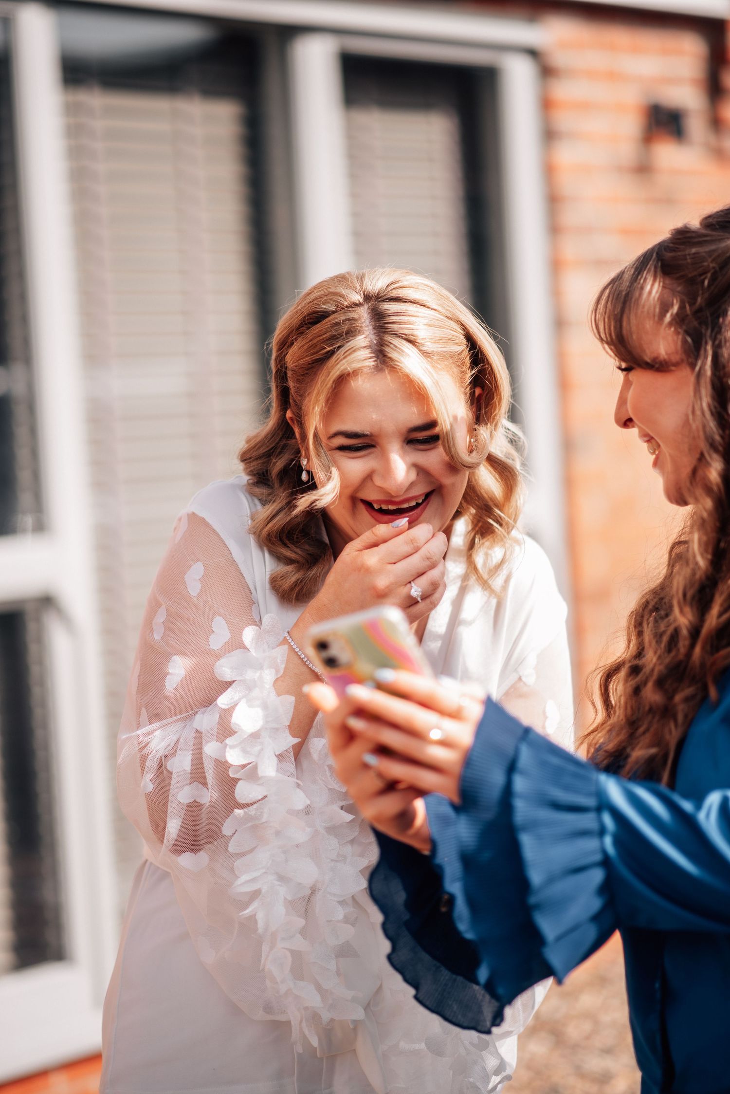 Laughing blonde bride in white floral gown sharing joyful moment with friend viewing phone outdoors.