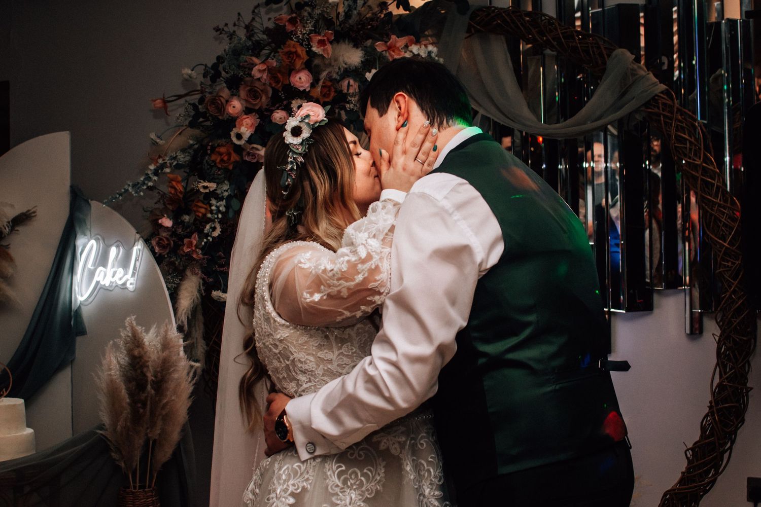 Bride and groom share first kiss under floral arch with neon sign at wedding.