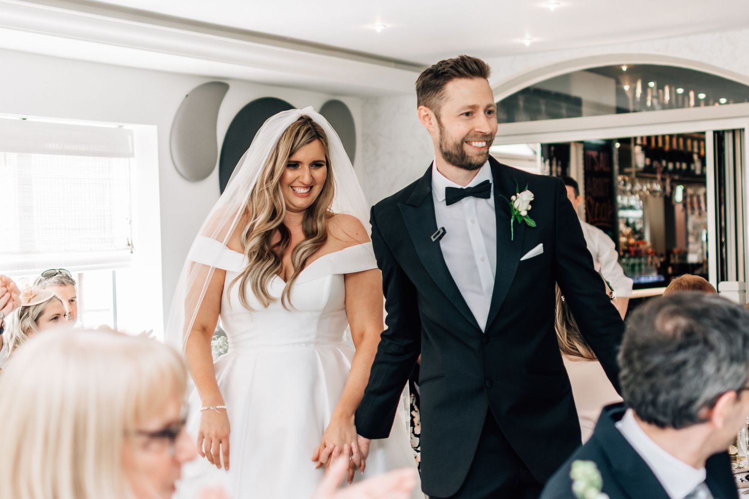 Bride and groom smiling and holding hands at wedding reception, bride in off-shoulder gown, groom in black tuxedo.