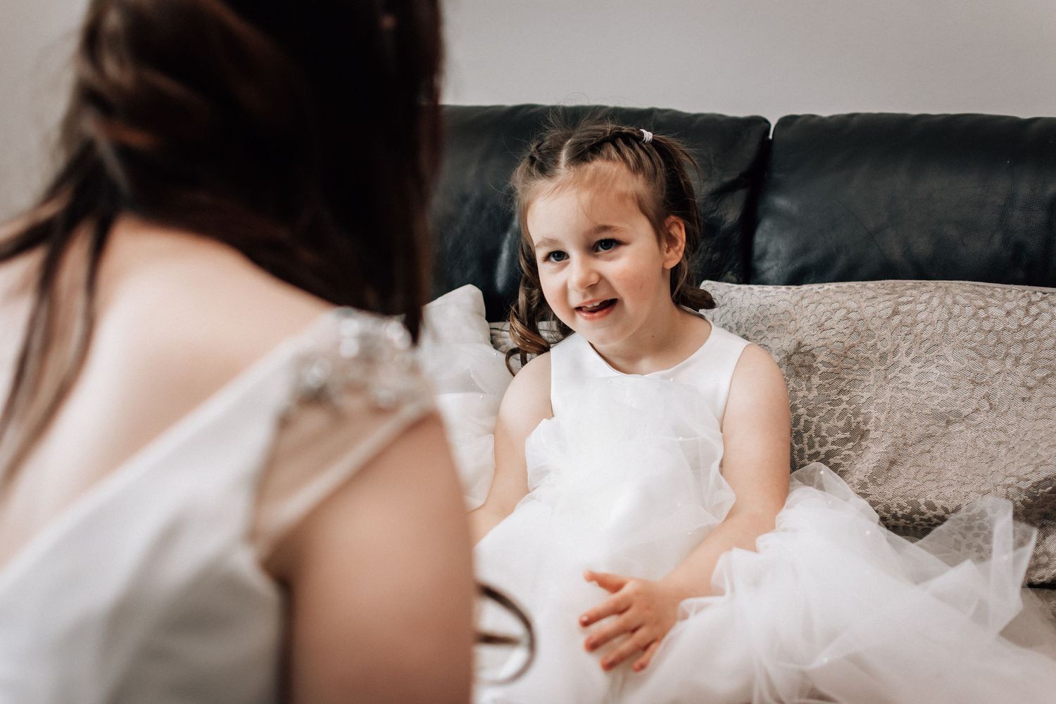 A smiling flower girl in a white tulle dress sits on a sofa, looking up at a bride during wedding preparations.