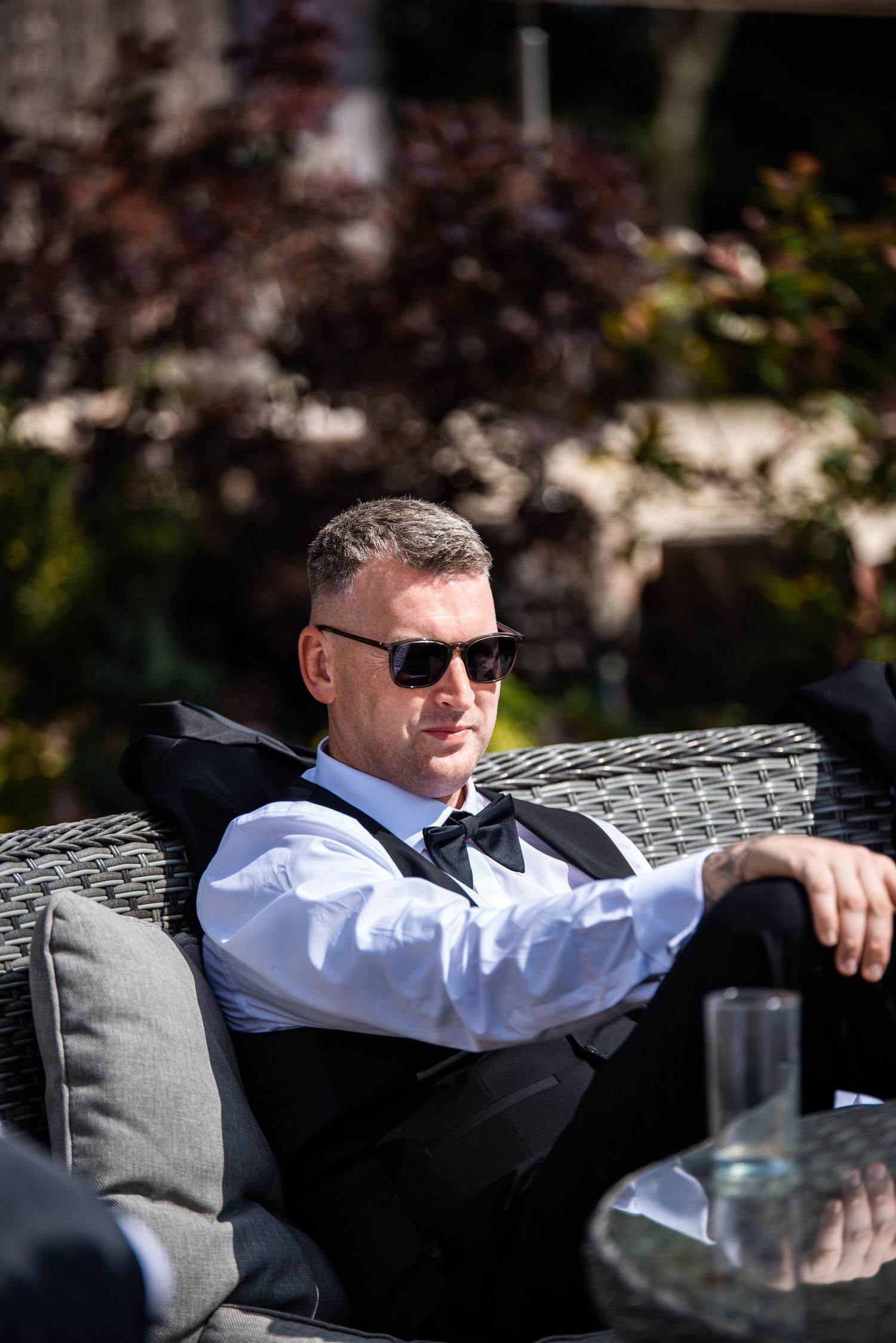A stylish man in a black vest and bow tie relaxes on outdoor furniture wearing sunglasses on a sunny day.