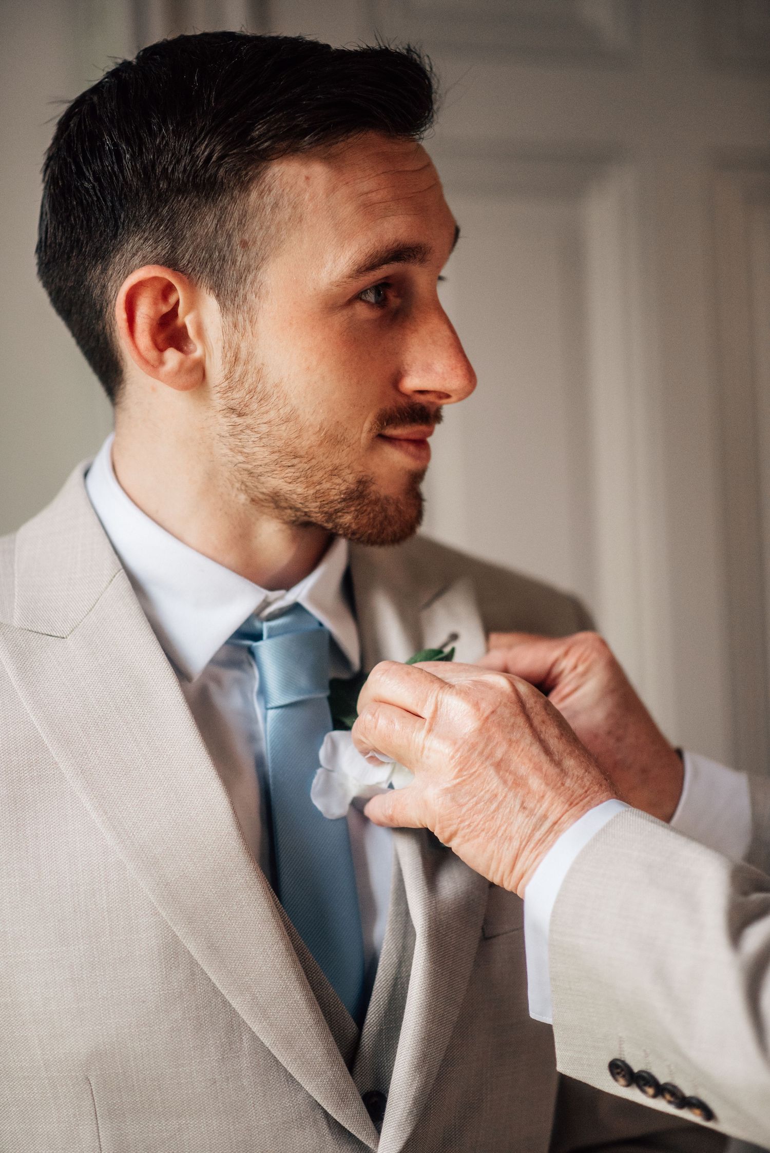 Groom in grey suit with blue tie having boutonniere pinned on lapel before wedding ceremony.