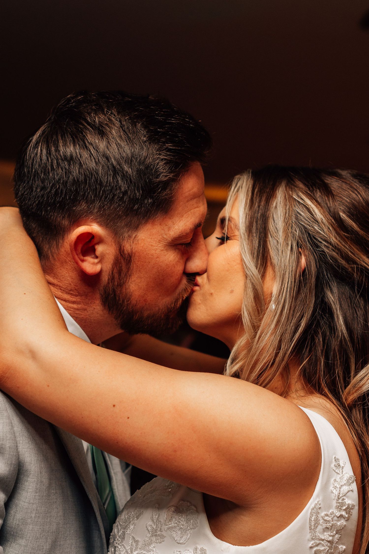 A couple shares a romantic kiss, with the bride in a white lace dress and groom in a gray suit.