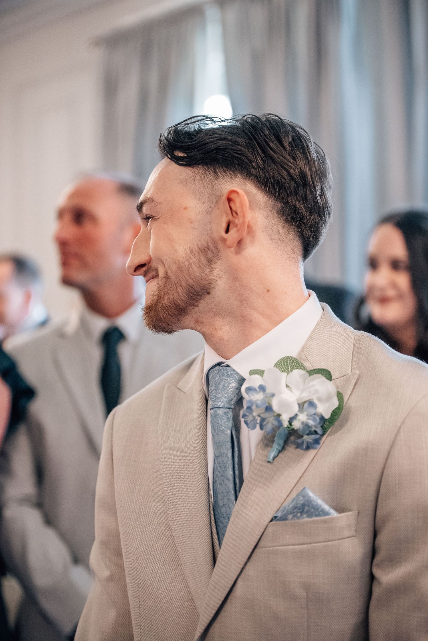 Smiling groom in beige suit with blue tie and floral boutonniere at wedding ceremony.