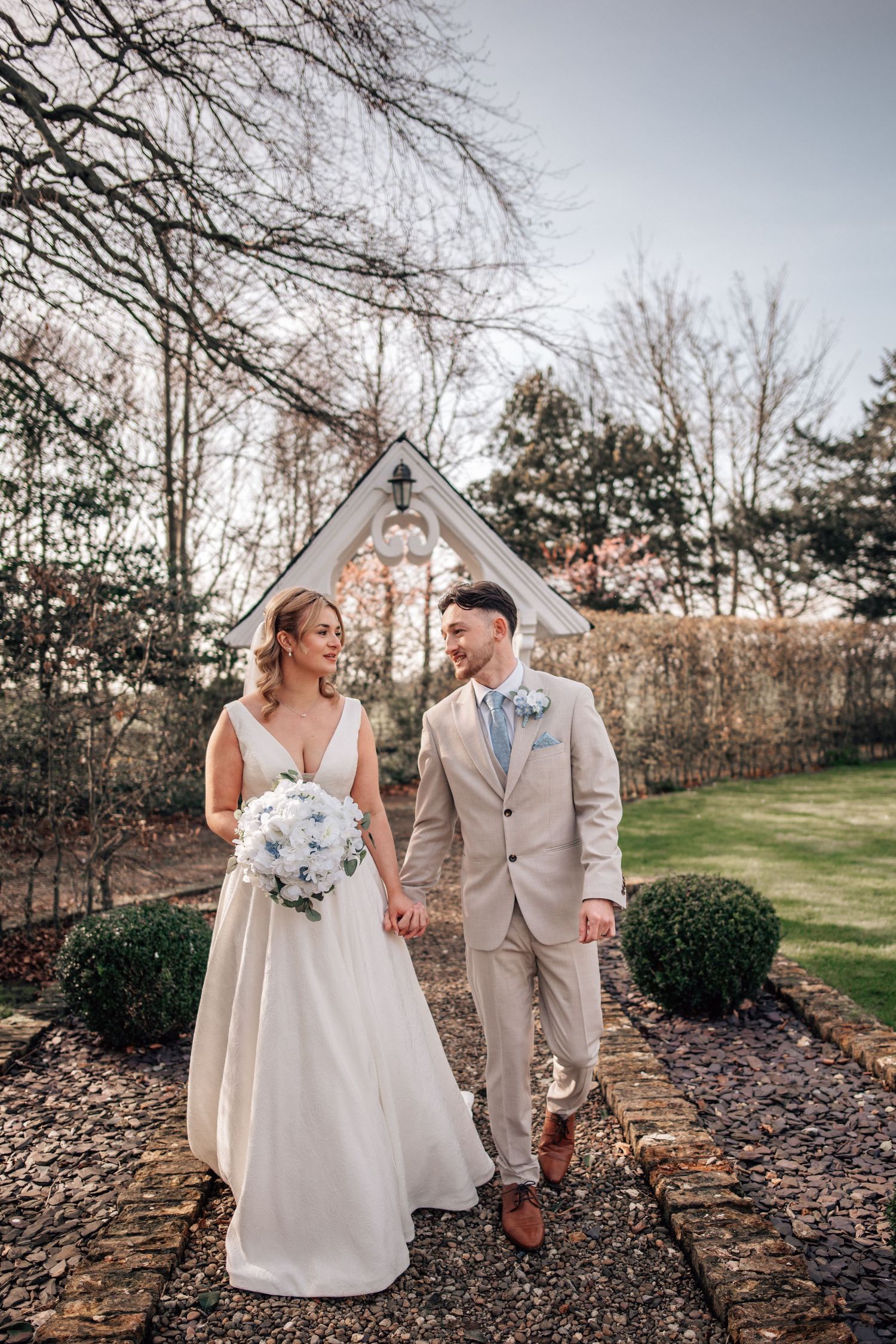Bride and groom walk hand-in-hand through a garden path, bride holding white bouquet, groom in beige suit.