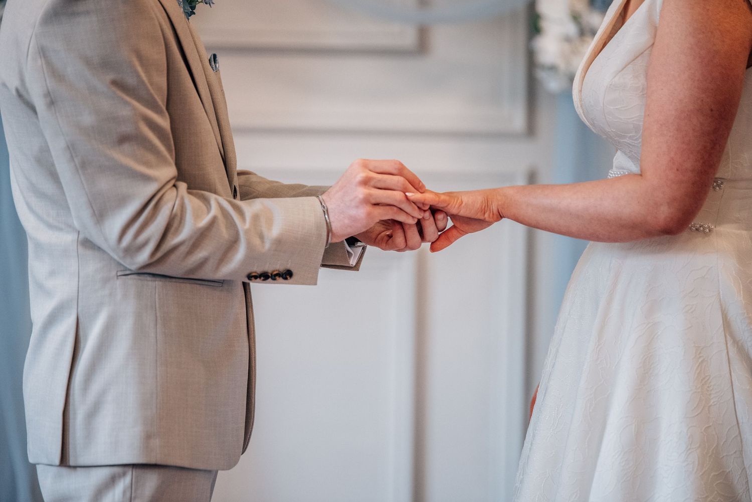 Groom in beige suit placing wedding ring on bride's finger during ceremony.