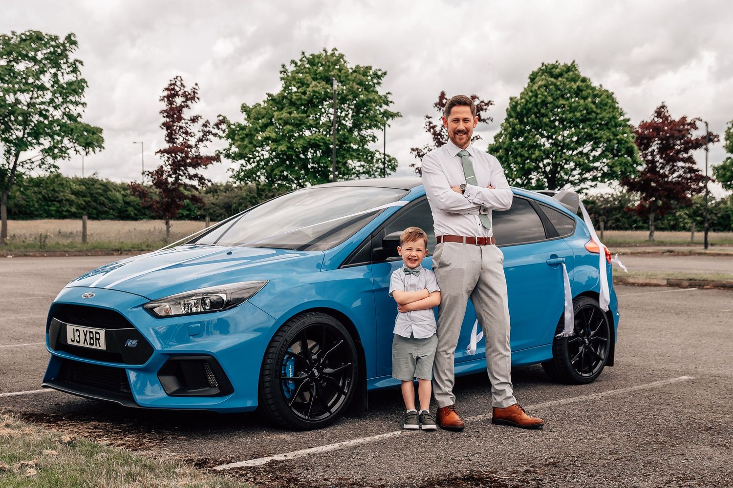 Man and young boy posing proudly in front of a blue Ford Focus RS in a parking lot.