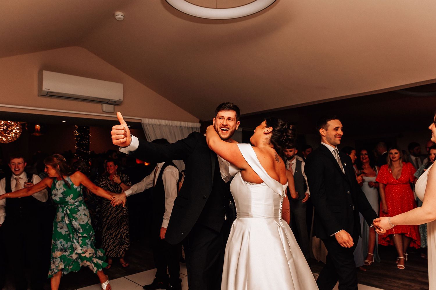 Bride and groom dancing joyfully with guests at a lively wedding reception, groom giving thumbs up.