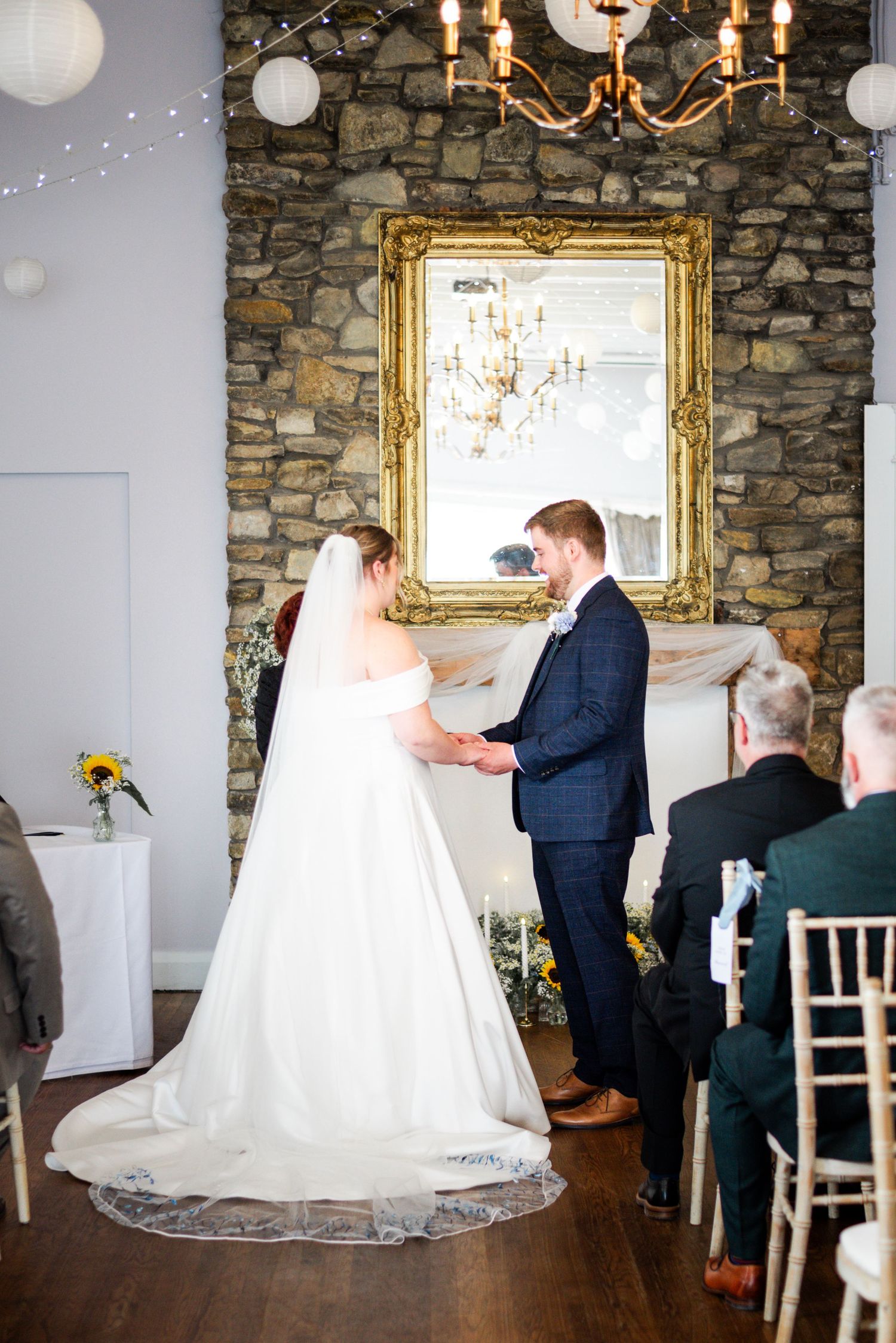 Bride and groom exchange vows at indoor wedding ceremony with stone wall backdrop and ornate gold mirror.