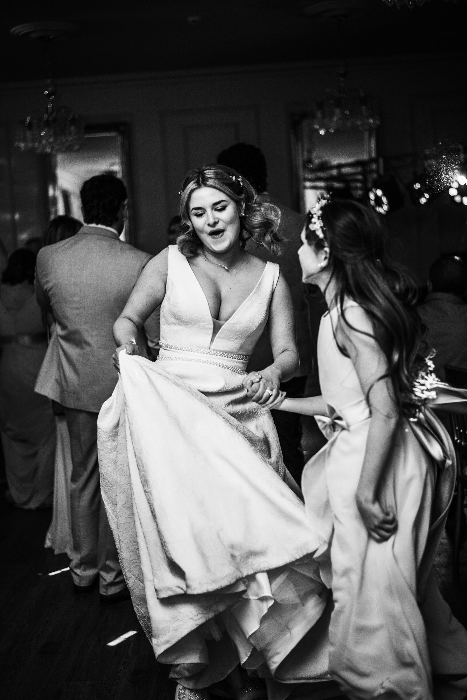 Bride laughing and dancing with bridesmaid at wedding reception, lifting white gown, black and white photo.